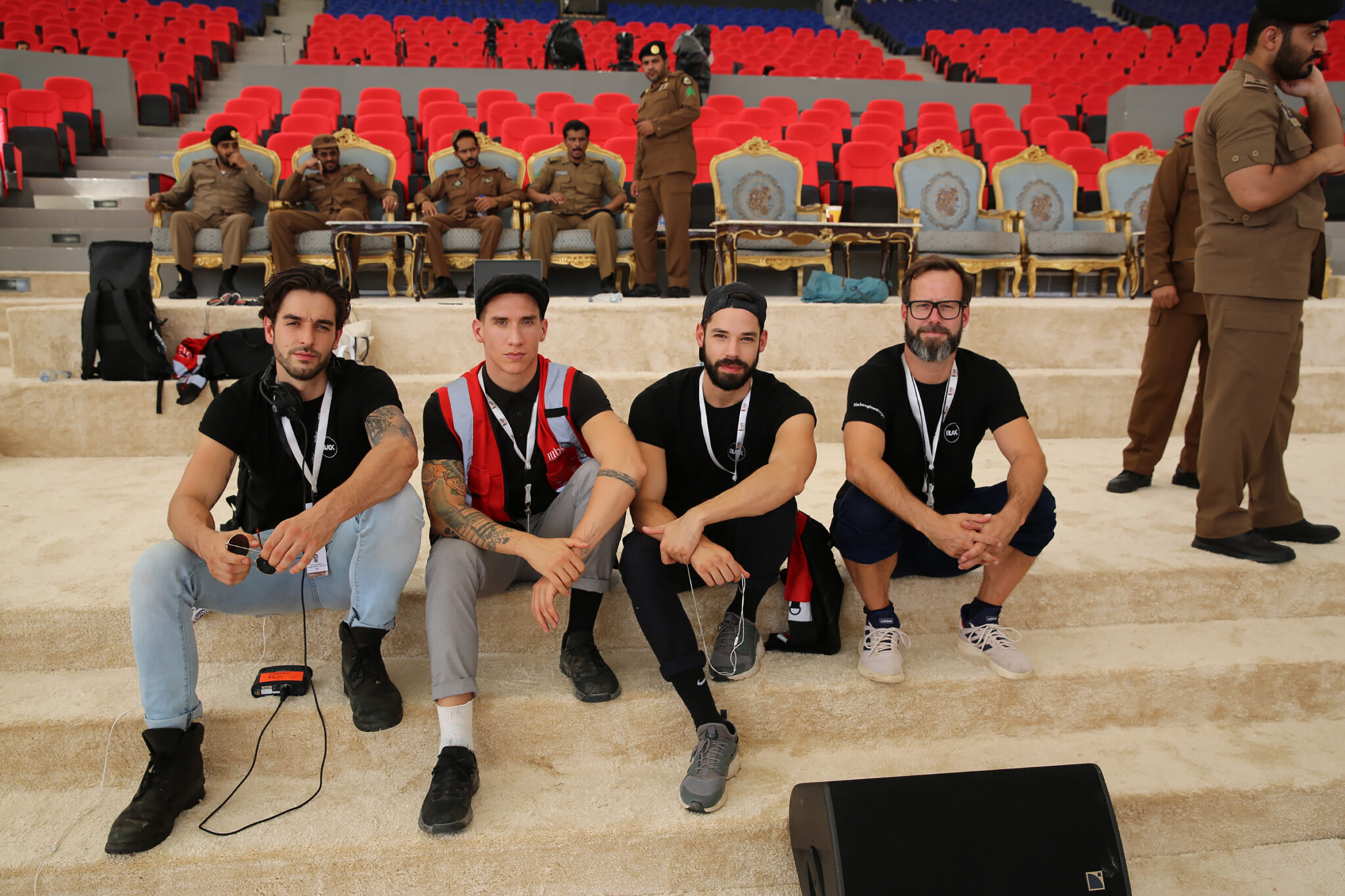 Five people in black shirts seated together in a row of red and white chairs, photographed during a break.