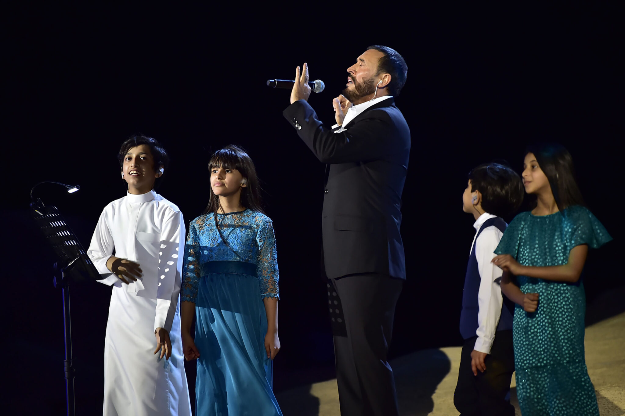 A singer in a black suit performing into a microphone on a dark stage, with four children in formal dress standing beside him.