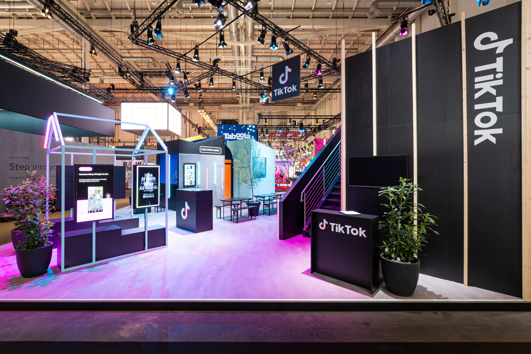 The corner of an exhibition stand with a tall dark wall, a vertical illuminated sign, an open staircase with a metal handrail, and a counter with potted plants on a pink-lit floor.