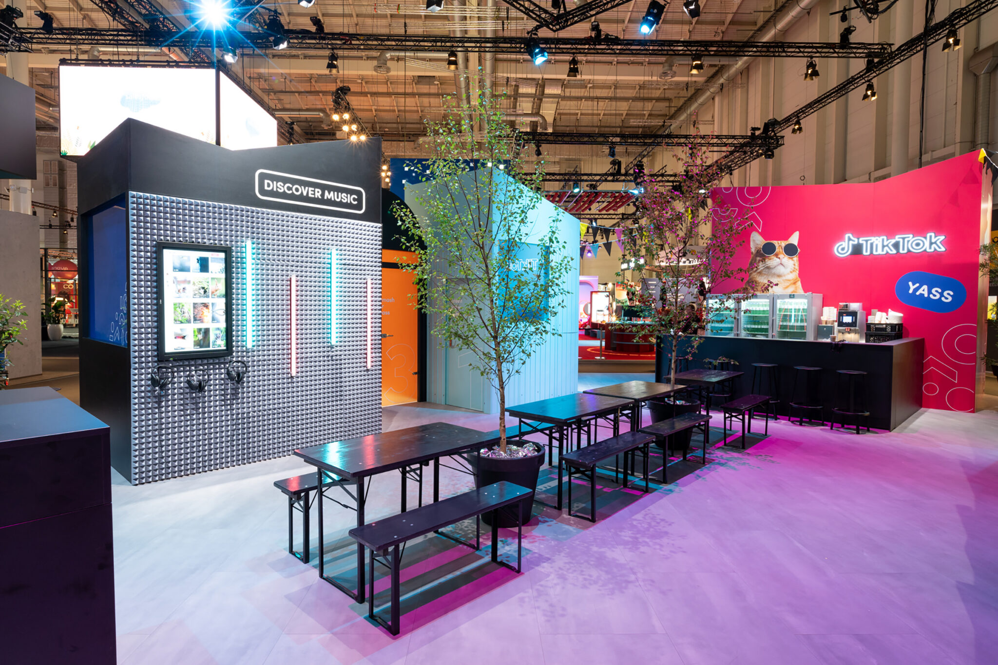 An exhibition seating area with long dark trestle tables and benches under small trees, facing a structure clad in silver studded panels, with a pink counter and bar stools to one side.