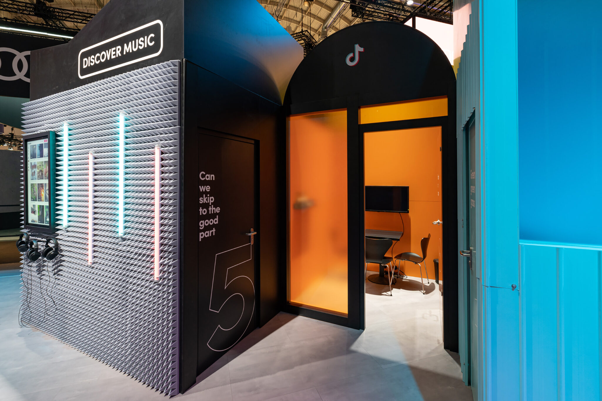 A small structure clad in grey acoustic foam panels with vertical neon tubes and hanging headphones, next to an arched black doorway opening onto an orange-walled room with a desk and two chairs.