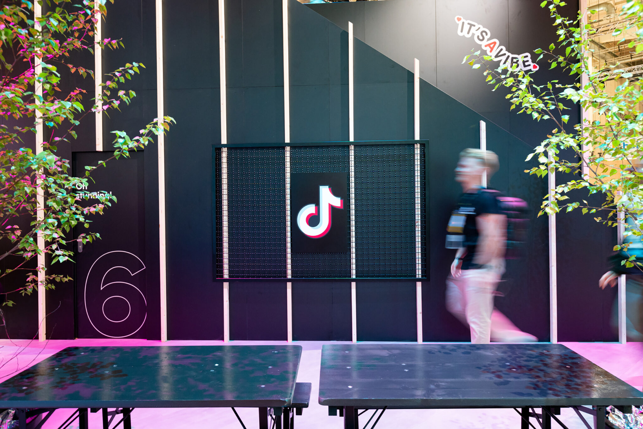 A dark slatted wall with a framed illuminated panel at its centre and a large outline numeral to one side, with a blurred figure walking past and leafy branches in the foreground.