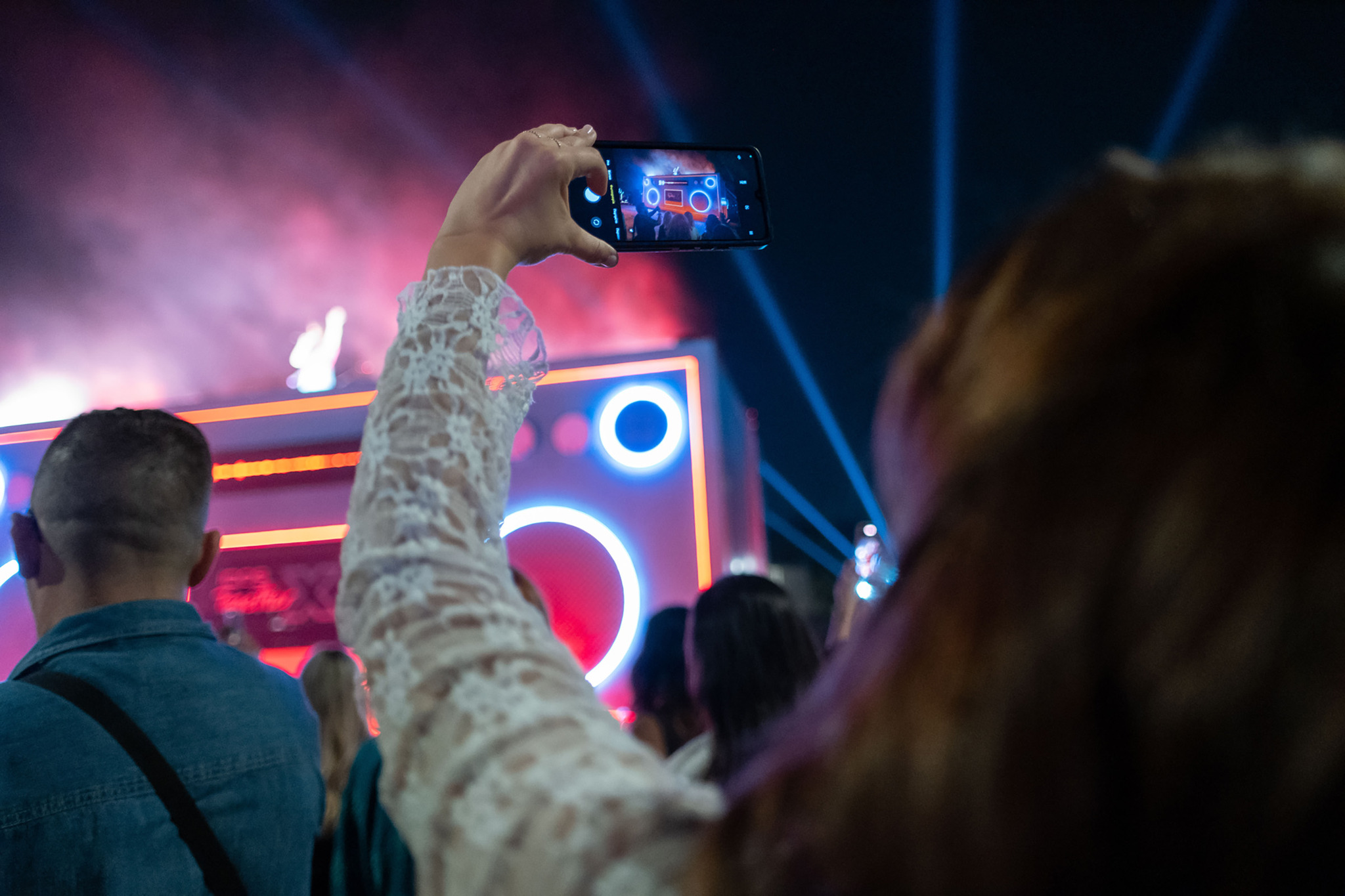 A woman films a stage shaped like a giant boombox with glowing circles on her phone, with blue light beams and a crowd around her.