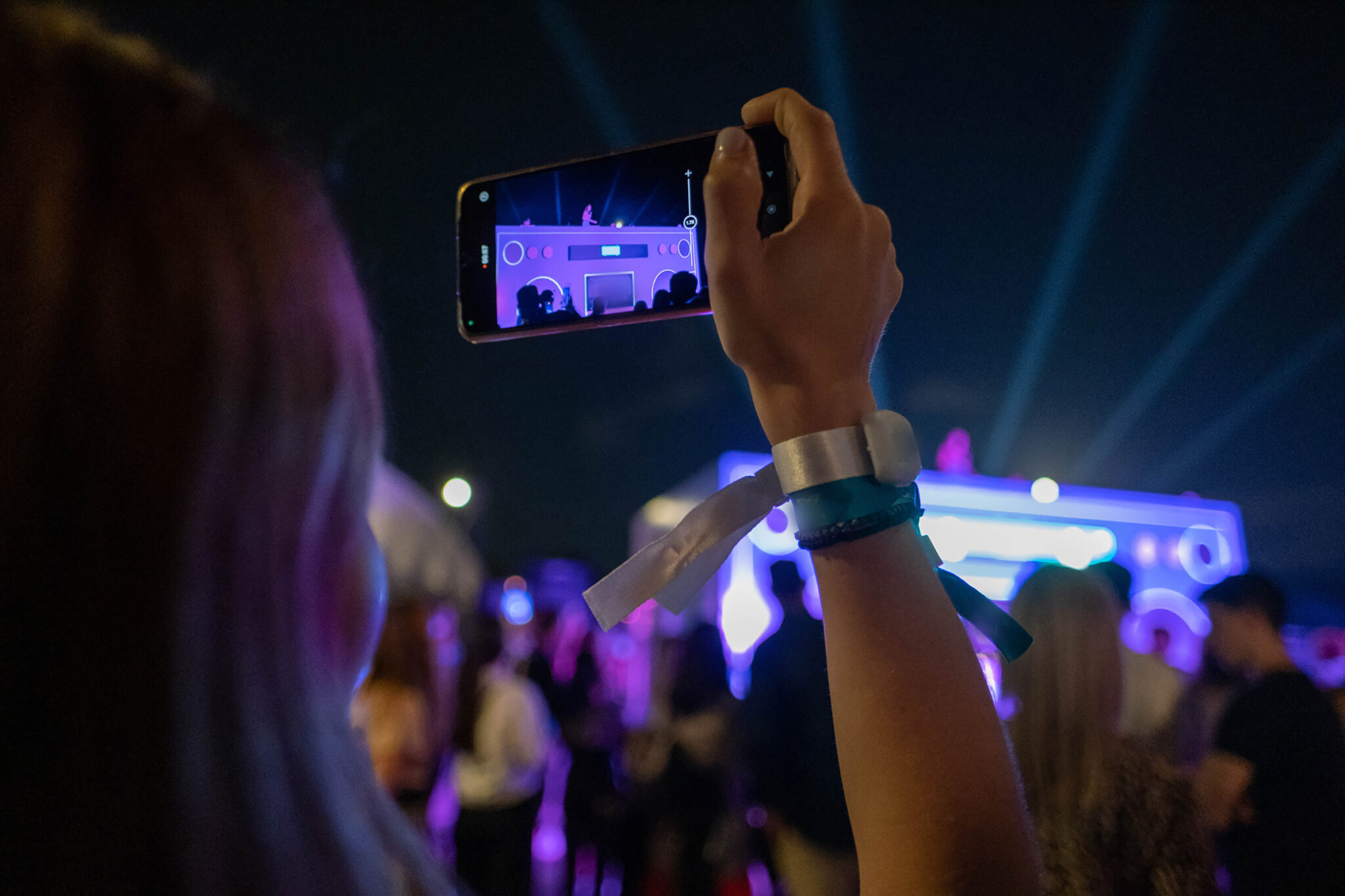 A woman in the audience holds up her phone to film a brightly lit stage at night, with blue light beams and a crowd in front of her.