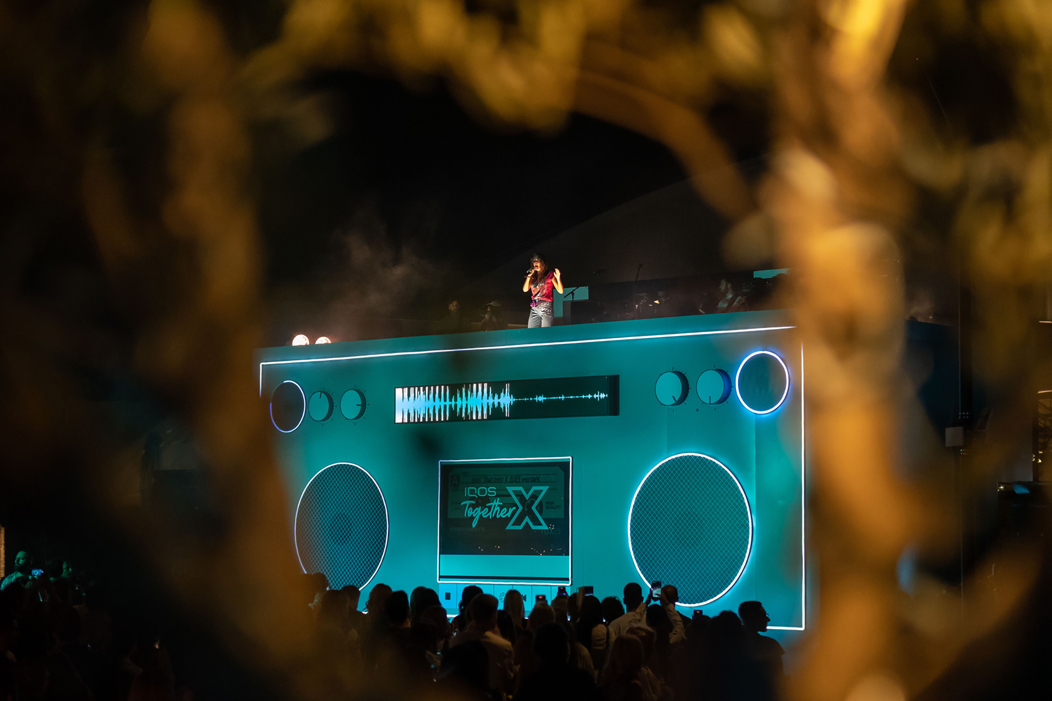 An outdoor stage built as a giant glowing boombox with round speakers and a sound wave display, a performer on top, seen through blurred lights in the foreground.