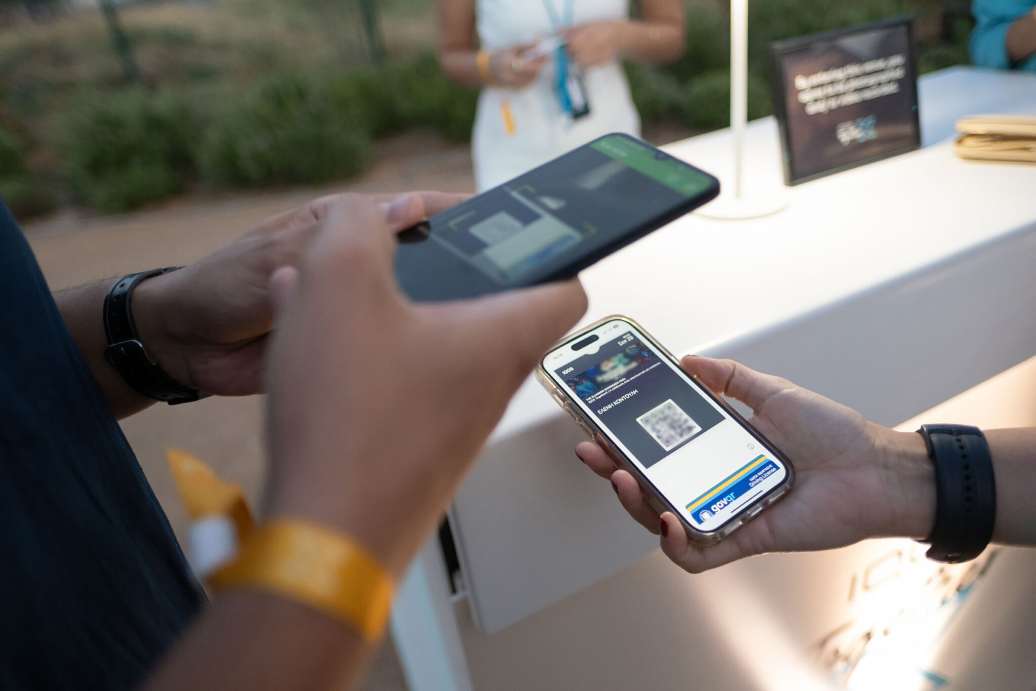 Close-up of hands holding two smartphones over a white counter, one showing a QR code, with a yellow wristband on one wrist.
