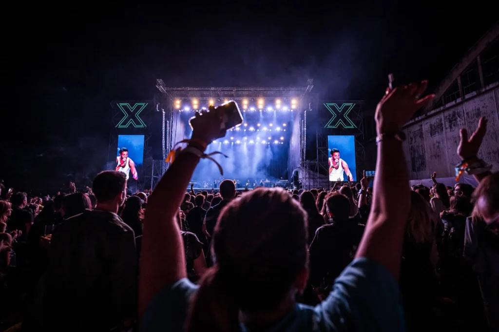 Concertgoers raise their arms and phones toward an outdoor stage with two large screens and glowing signs on either side.