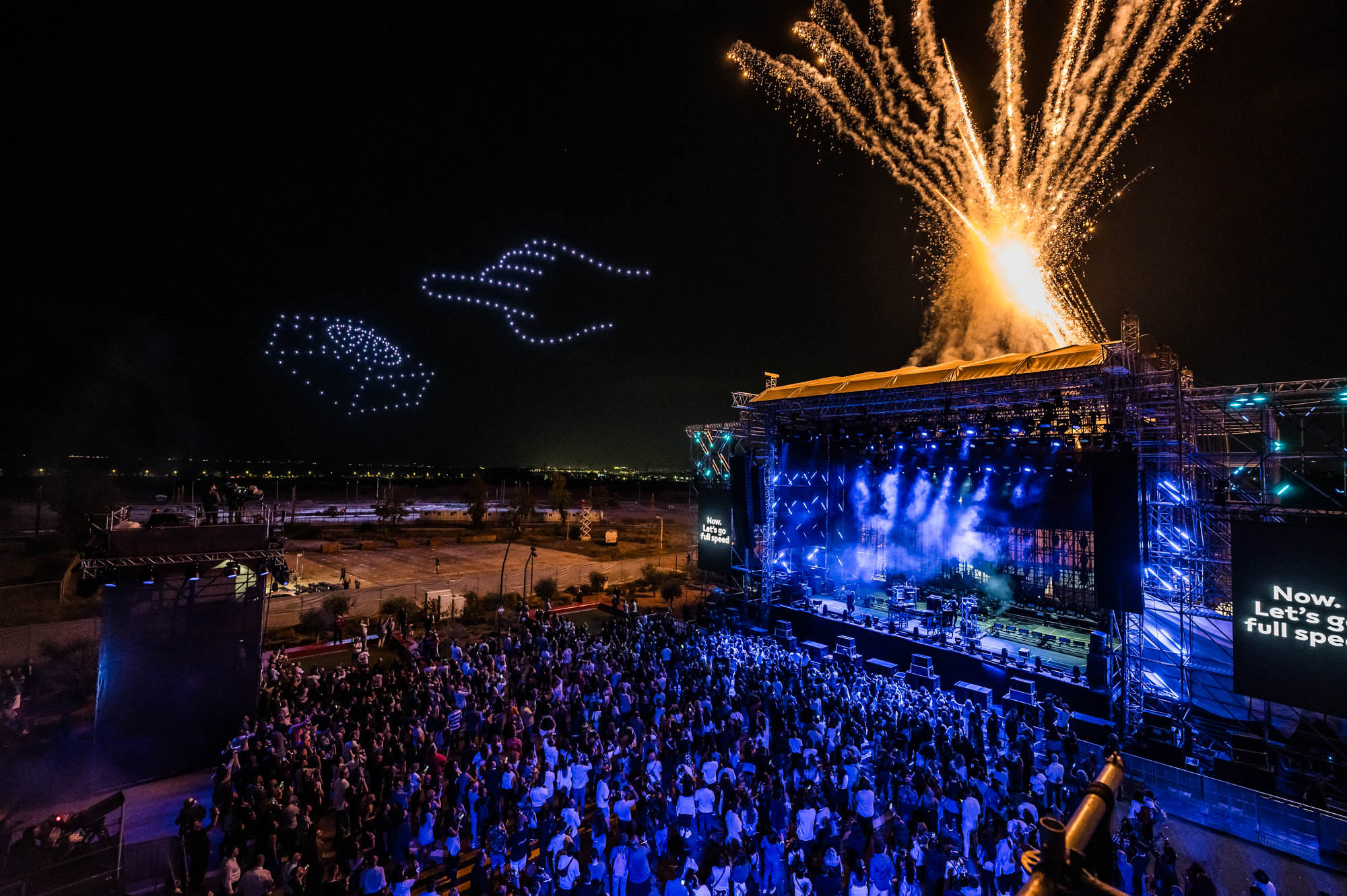 Drones form glowing shapes in the night sky while fireworks burst from the roof of an outdoor stage above a large crowd.