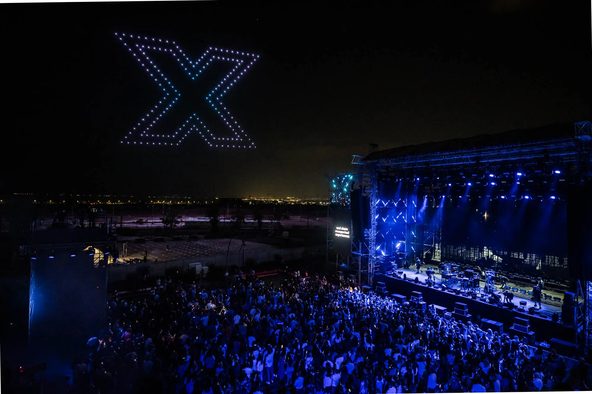 Hundreds of lit drones form a large X shape in the night sky above an open-air concert stage lit in blue, with a large crowd below.