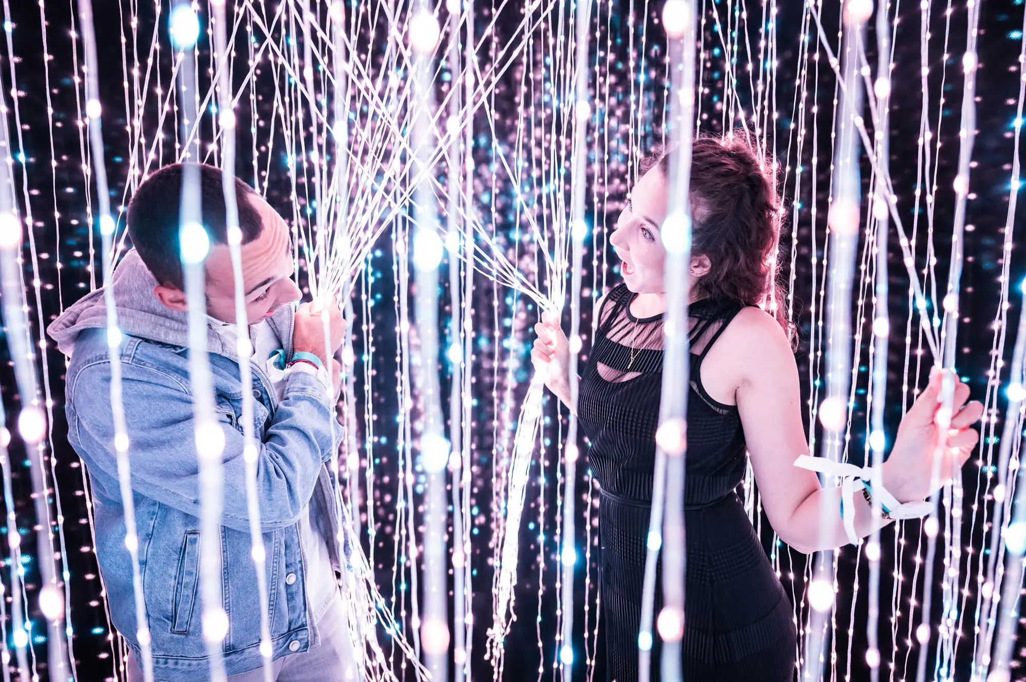 Two people stand inside a curtain of hanging light strings, one parting the strands with their hands, surrounded by small white lights.