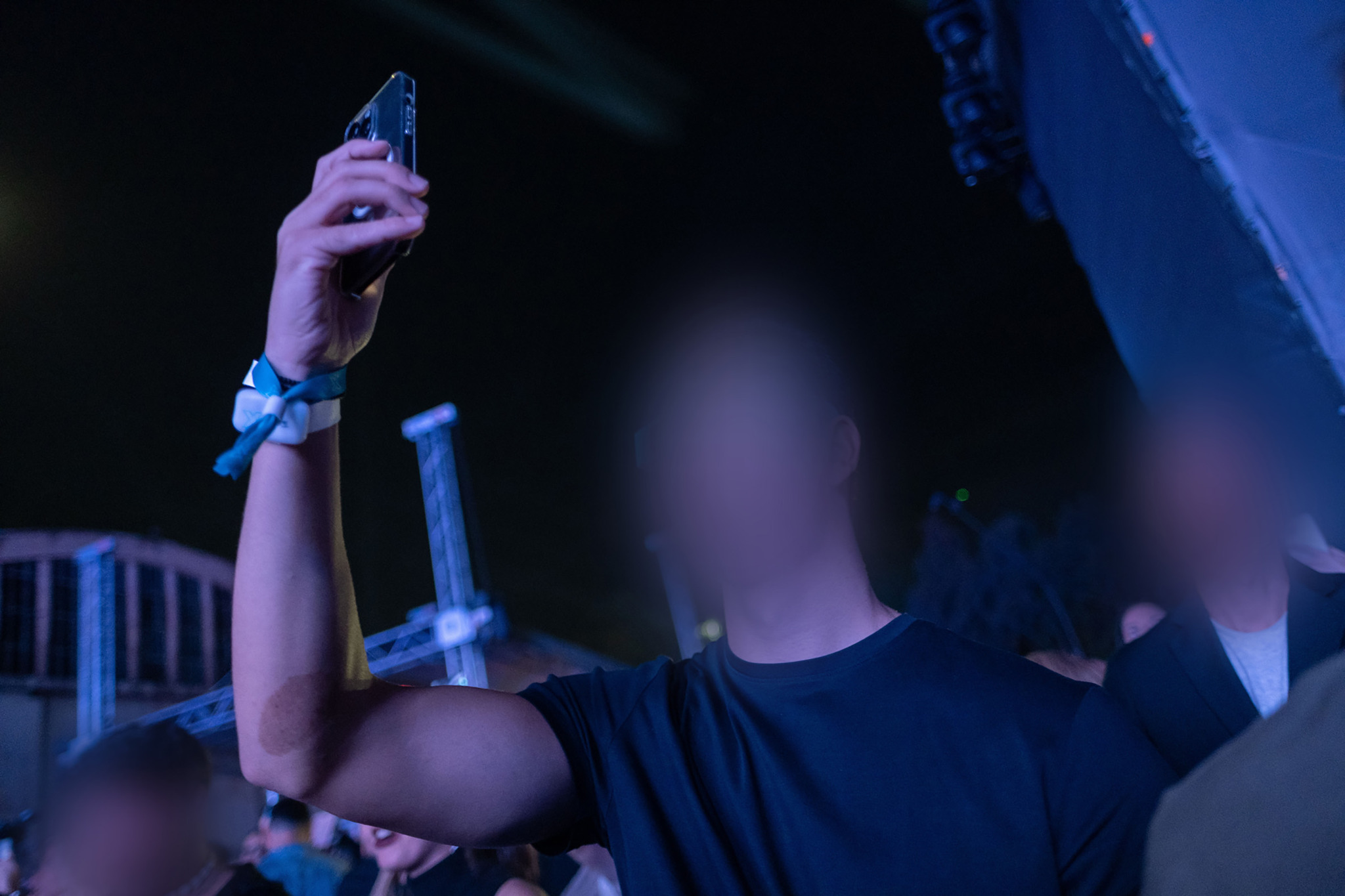 A raised arm wearing a wristband holds up a phone to take a photo in a crowd at night near a lit stage.