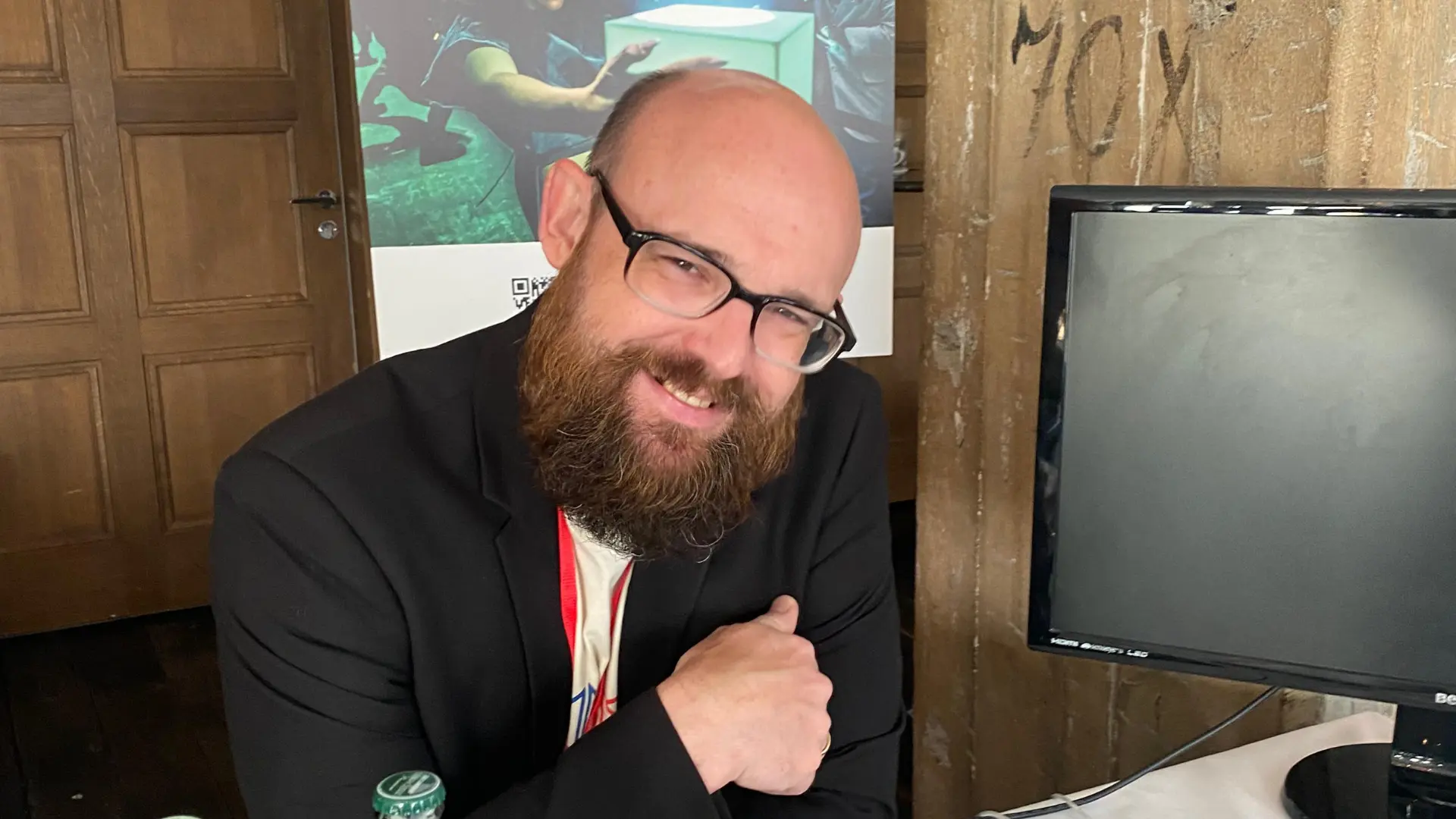 Jon Buckels smiles while sitting at a table, with a monitor and a poster in the background. Battle Royal Studios