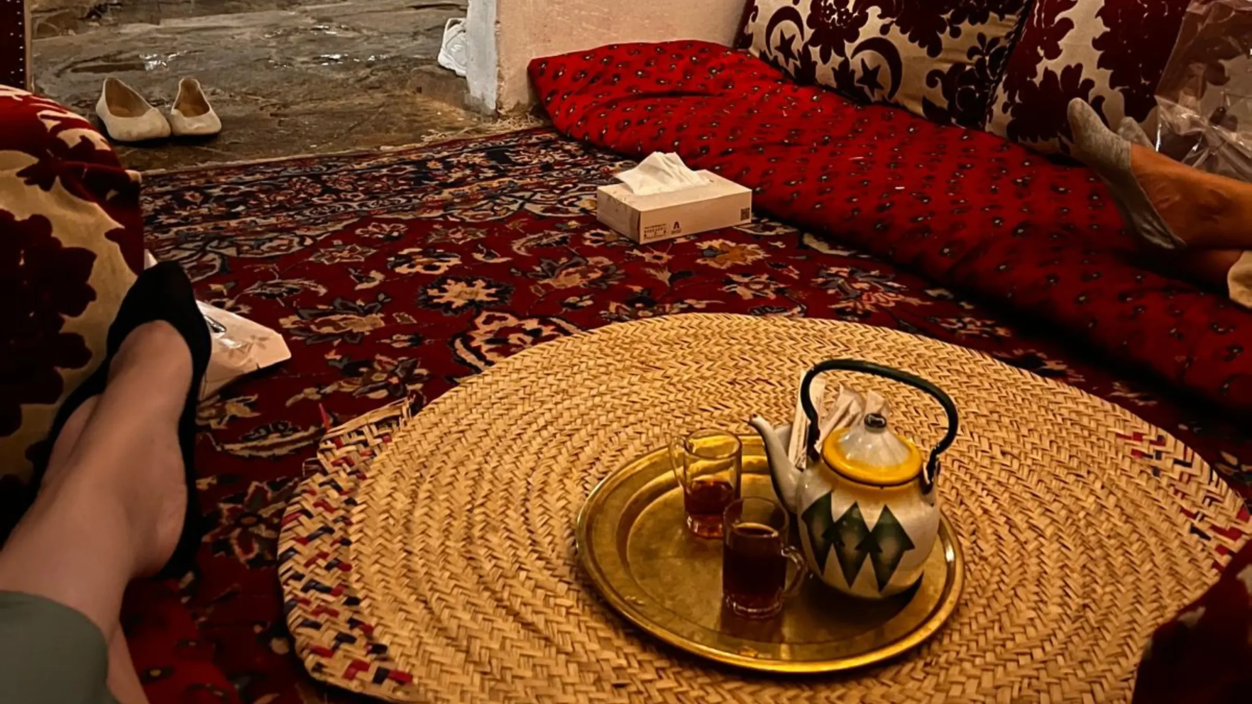 A cozy sitting area with a teapot and two glasses of tea on a tray, placed on a woven mat over a patterned rug. Cushions and a box of tissues are nearby. Battle Royal Studios