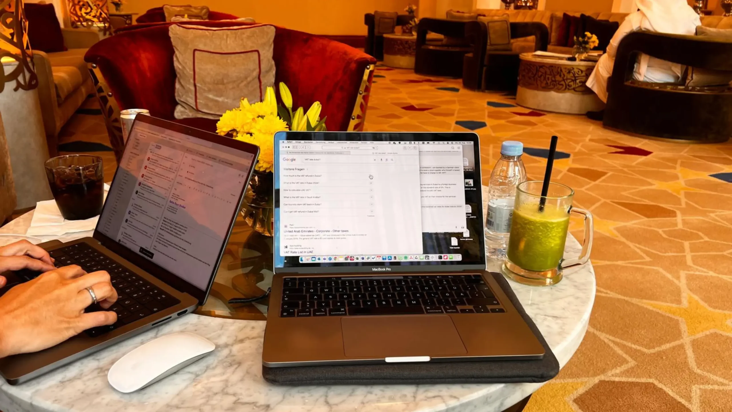 Two laptops are open on a marble table, with a person typing. Drinks, including a green juice and water bottle, are placed next to the computers in a comfortable lounge setting. Battle Royal Studios