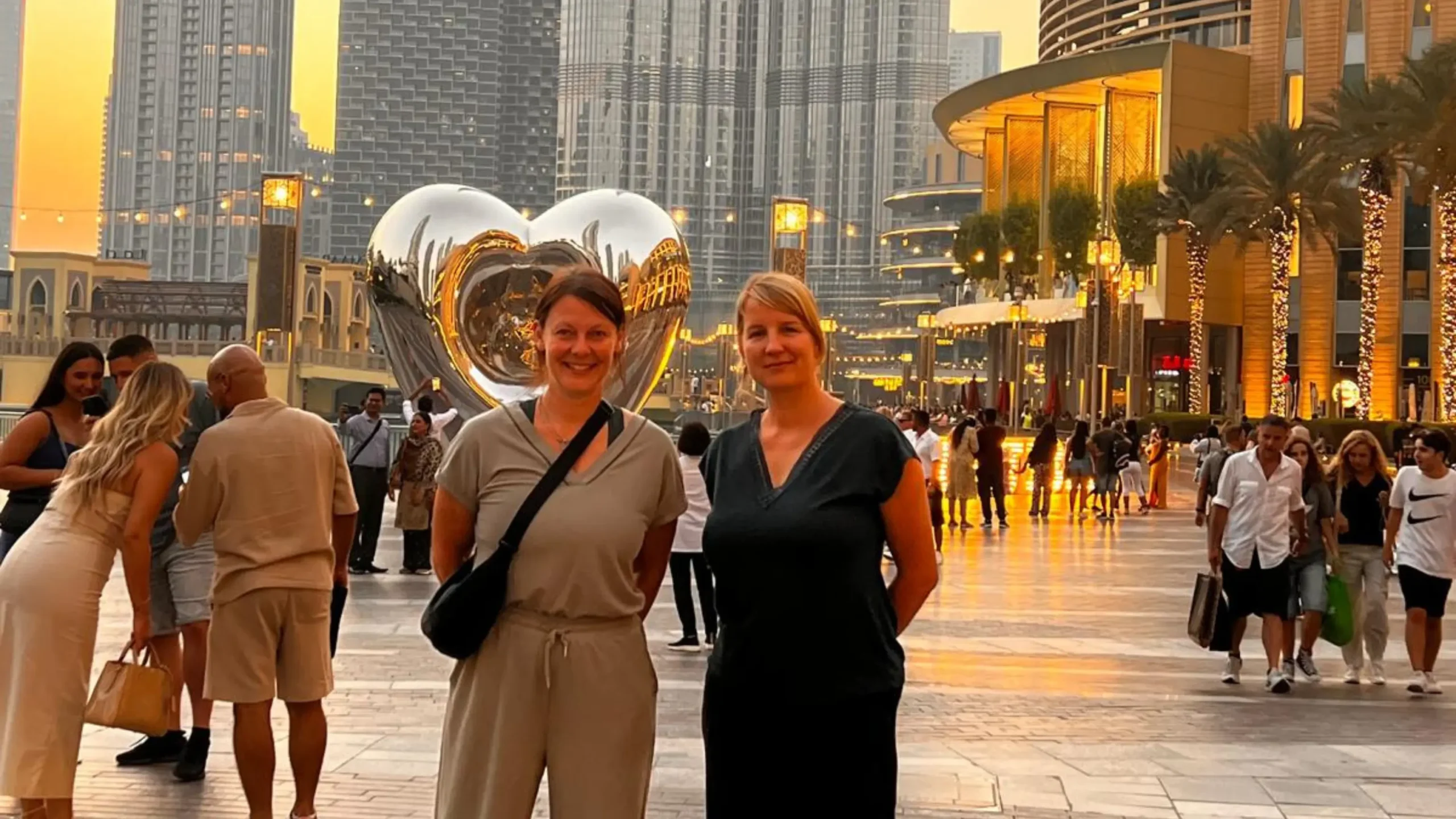 Sarah La Tragna and Judith Hoch stand smiling in front of a large metallic heart sculpture, with a busy city scene and skyscrapers in the background. Battle Royal Studios