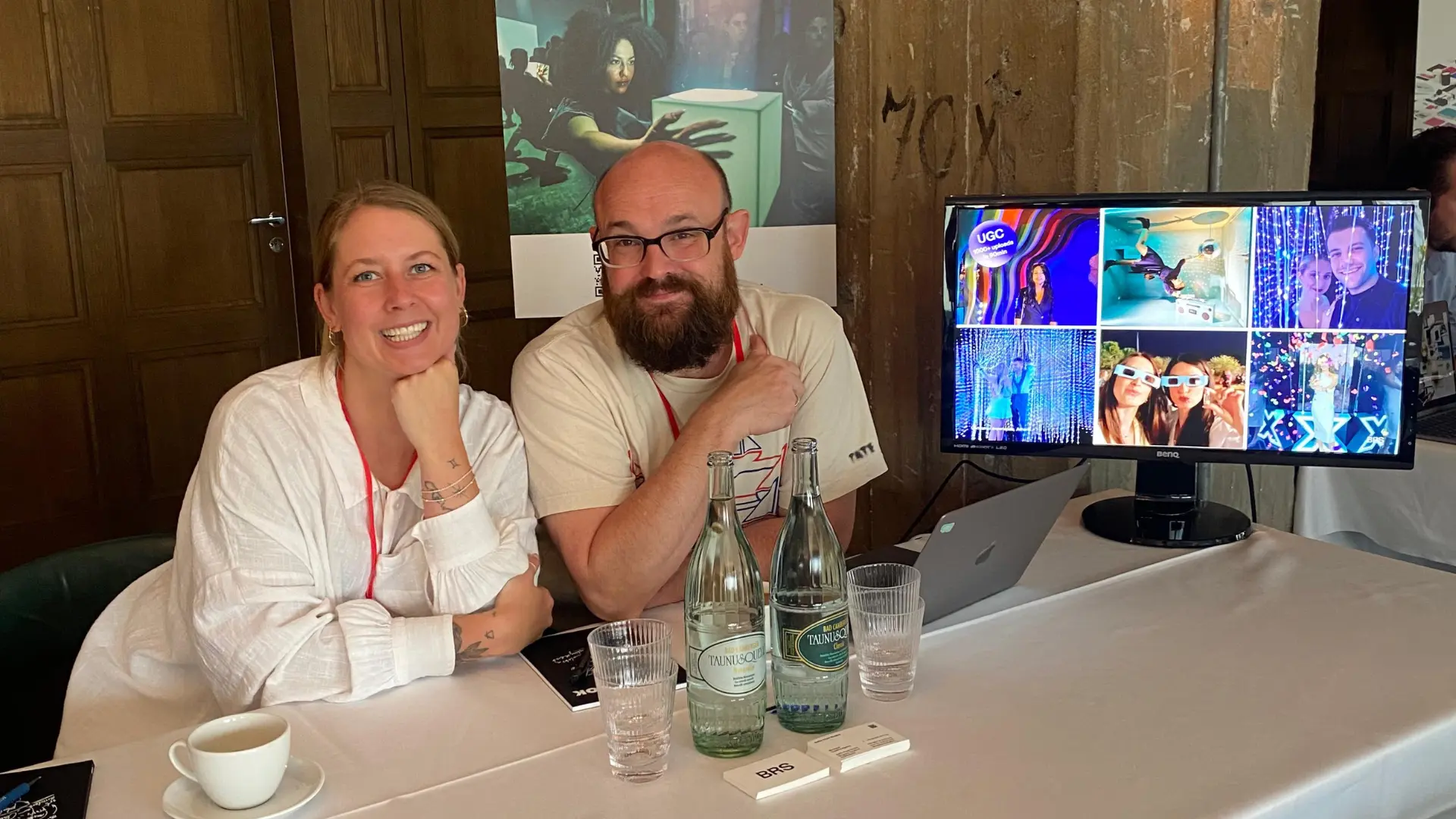 Janina Schaffrath and Jon Buckels smile while sitting at a table with water bottles and a monitor displaying vibrant images. Battle Royal Studios