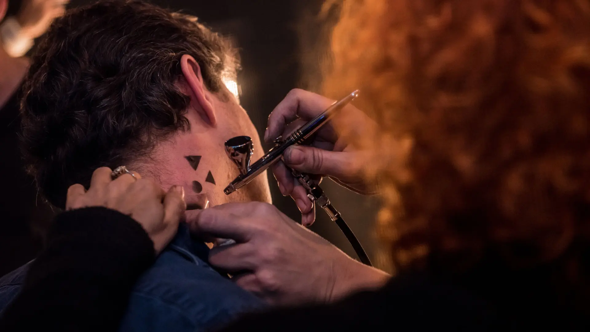 A person is getting a temporary tattoo airbrushed onto the side of their neck, with small triangular shapes being applied. Battle Royal Studios