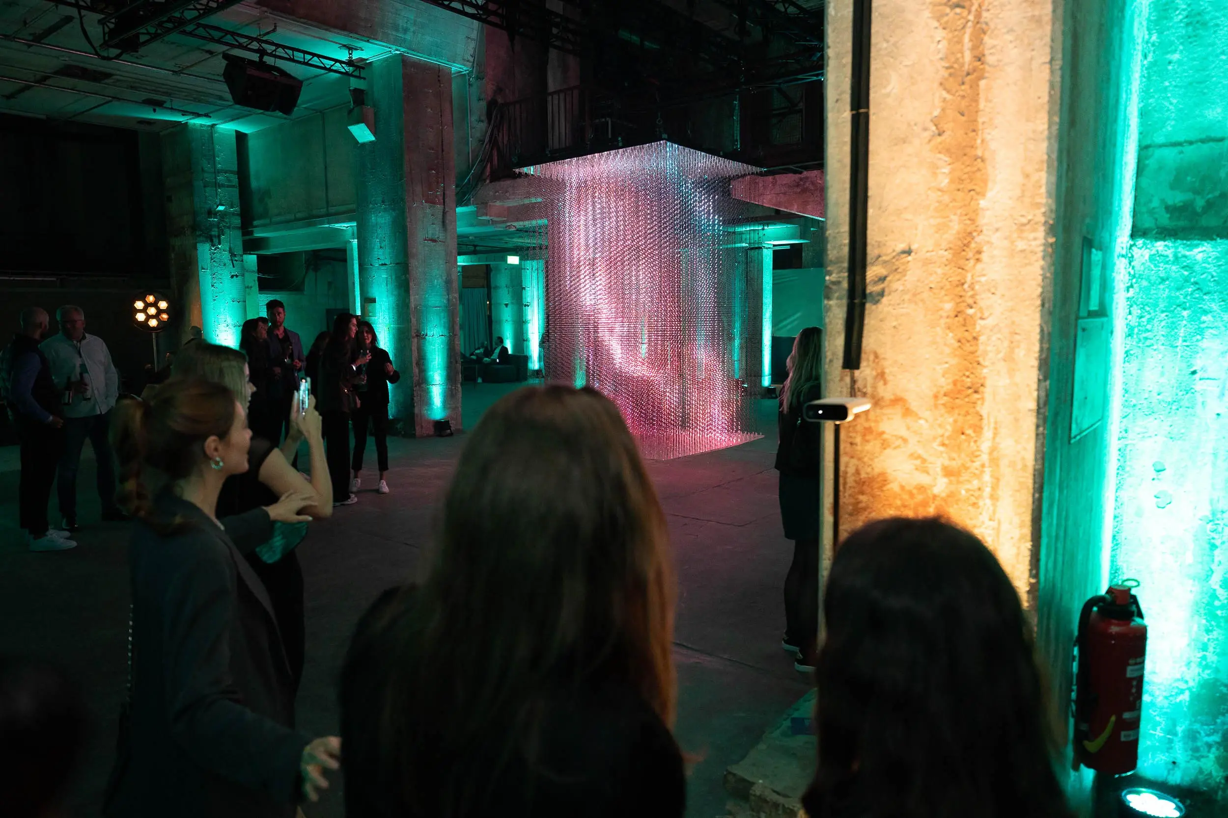 People in a teal-lit industrial room, watching a hanging art installation. Battle Royal Studios