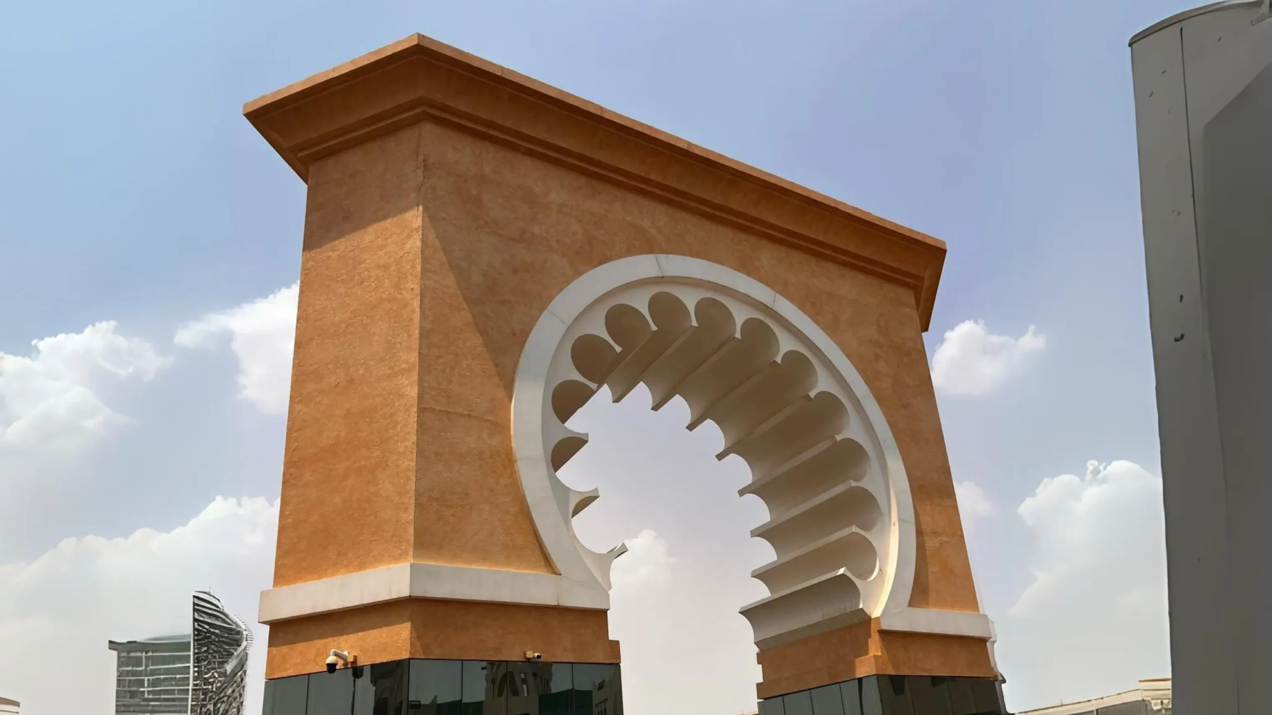 A large decorative archway with intricate design stands against a bright, cloudy sky. Modern buildings can be seen in the background. Battle Royal Studios