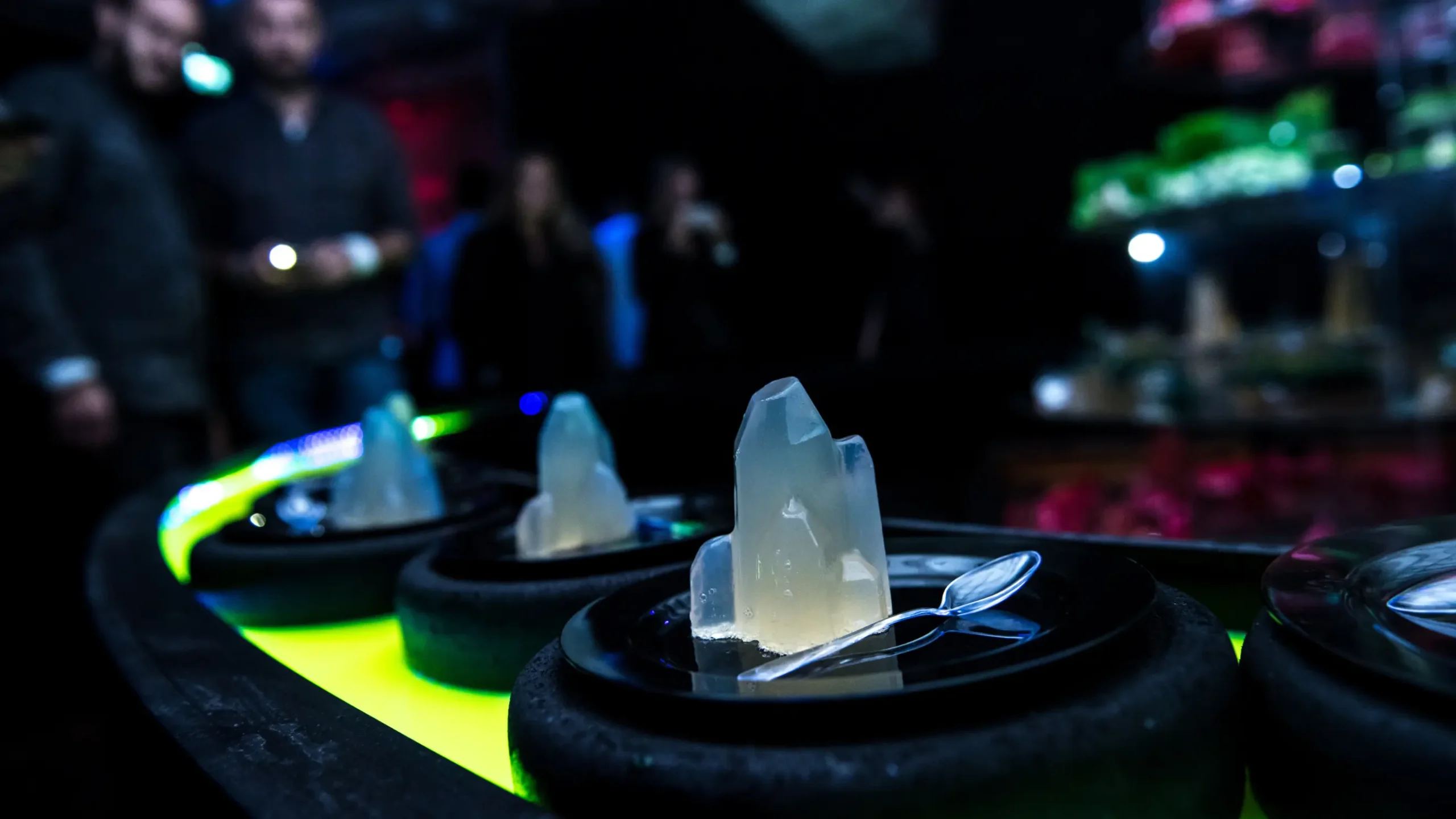 A display of artistic crystal-like desserts arranged on plates, with small spoons beside them, glowing under soft lighting in a dark setting. Battle Royal Studios