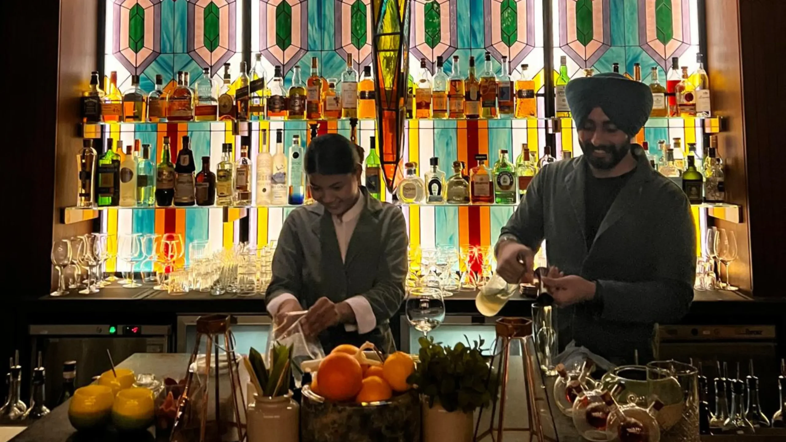 Two bartenders are preparing drinks behind a bar, with colorful stained glass and a variety of bottles displayed on shelves in the background. Battle Royal Studios