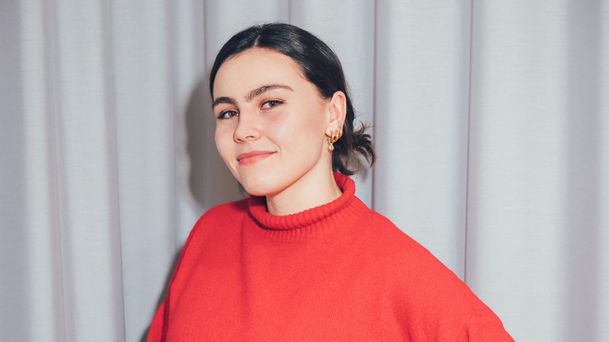 A young woman in a red jumper smiling in front of a pale curtain