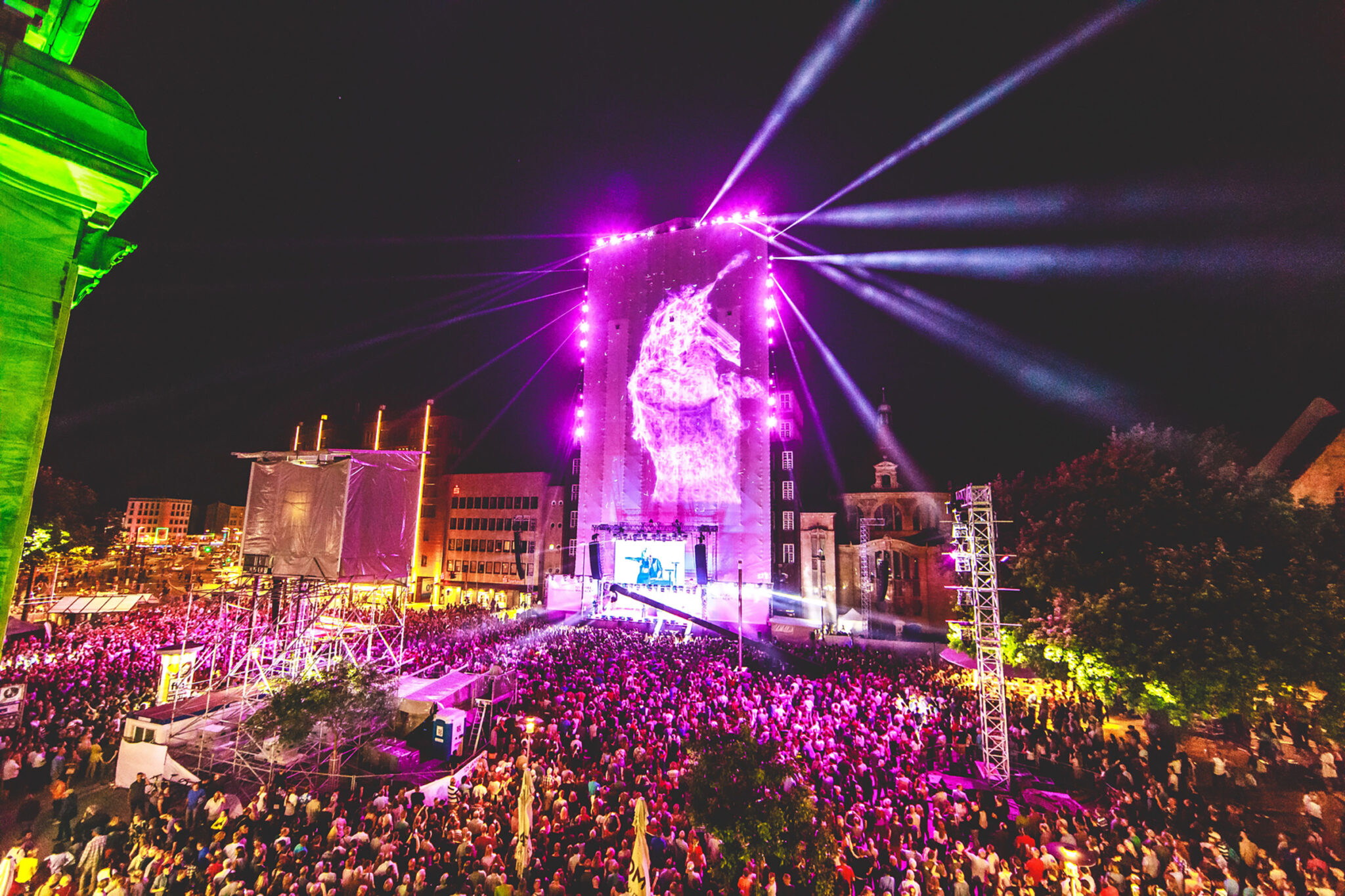 Night view across a packed city square, with a tall building lit in pink showing a flame-like projection and beams of light fanning out above the stage.