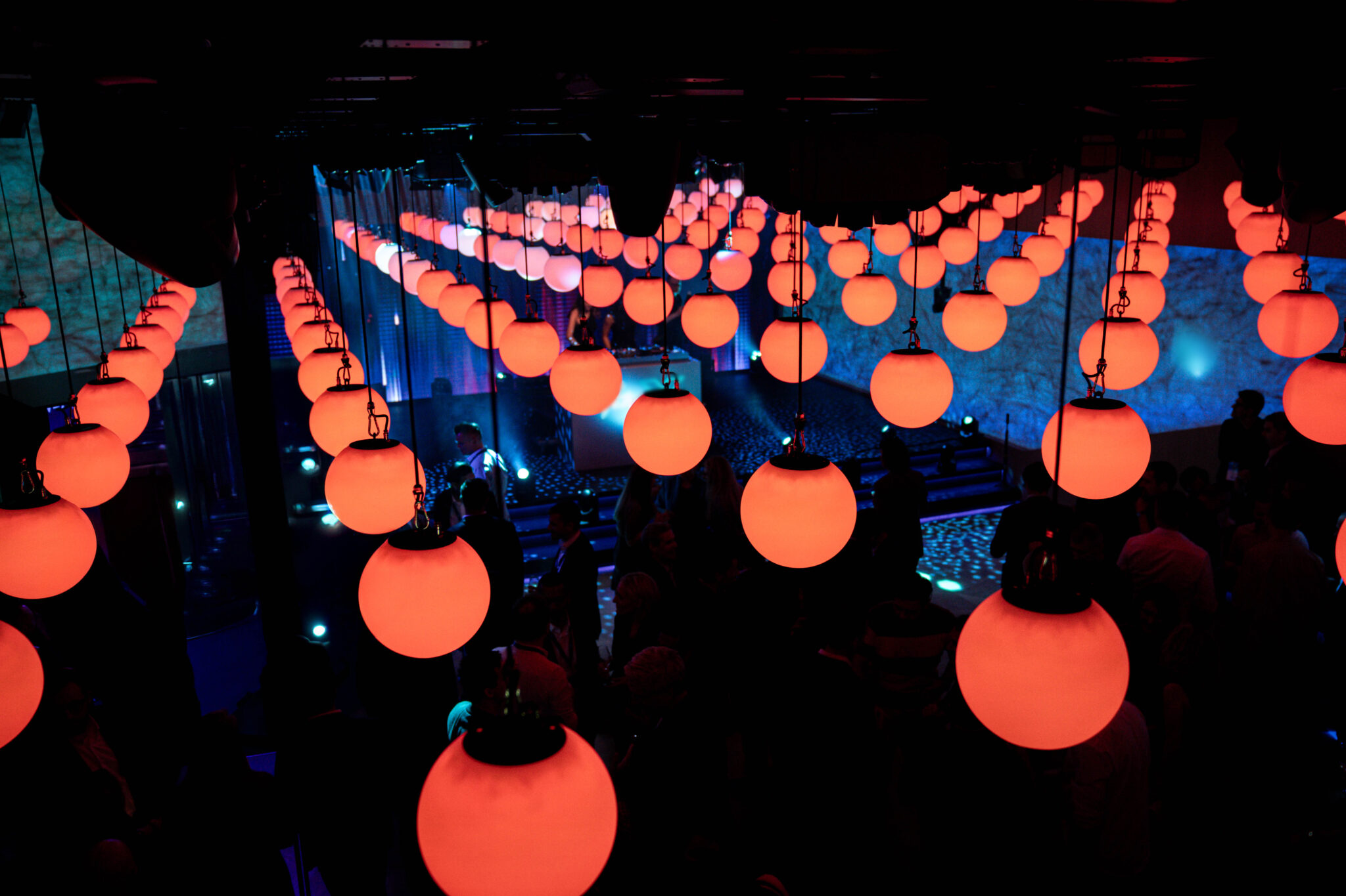 Large orange and white glowing spheres hanging in rows above a dark room full of people.