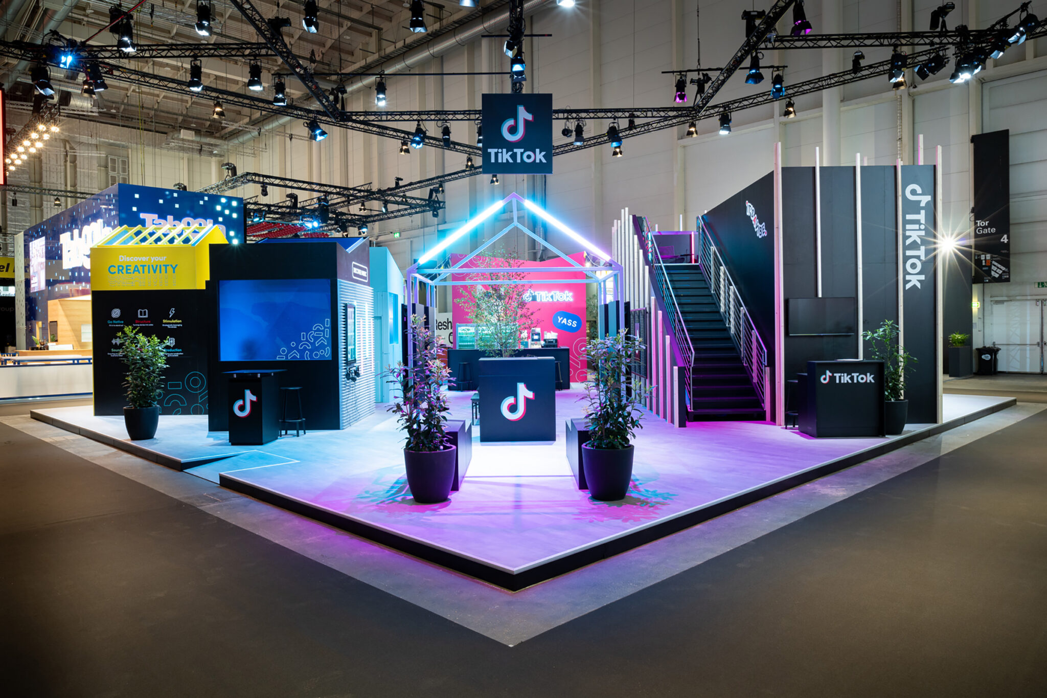 A trade fair stand built as a cluster of small house-shaped structures on a pale raised platform, lit in pink and blue, with an open staircase at one side and potted shrubs along the front edge.