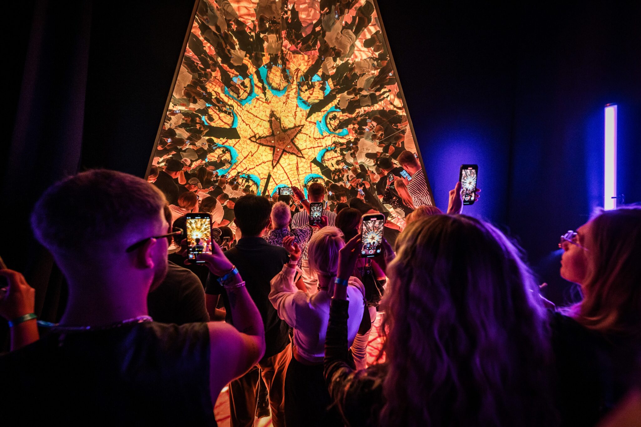 Guests photographing a glowing kaleidoscope-style light installation with their phones in a dark room.