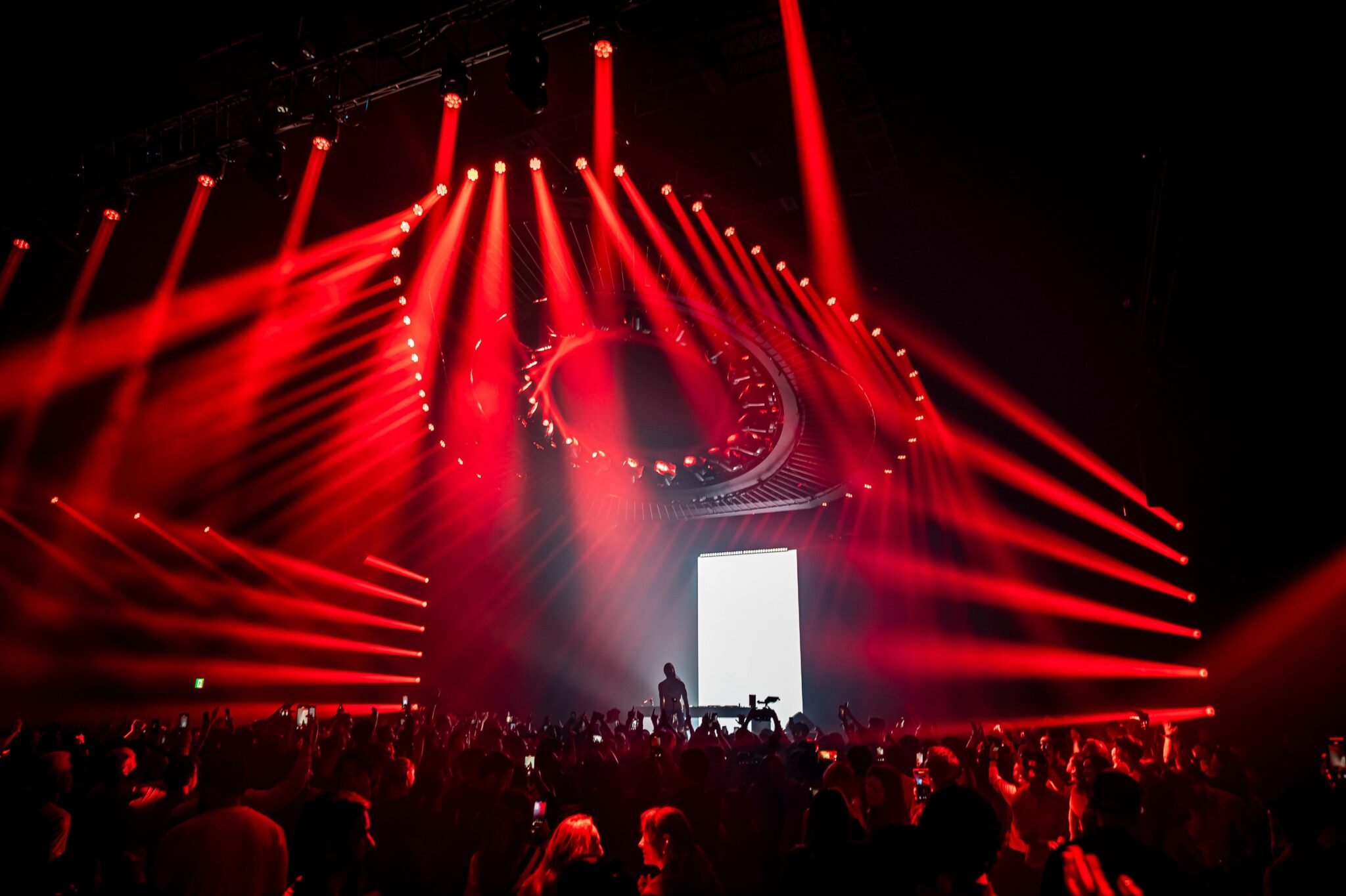 Red laser beams fanning out over a dark stage with a bright vertical screen and a silhouetted audience.