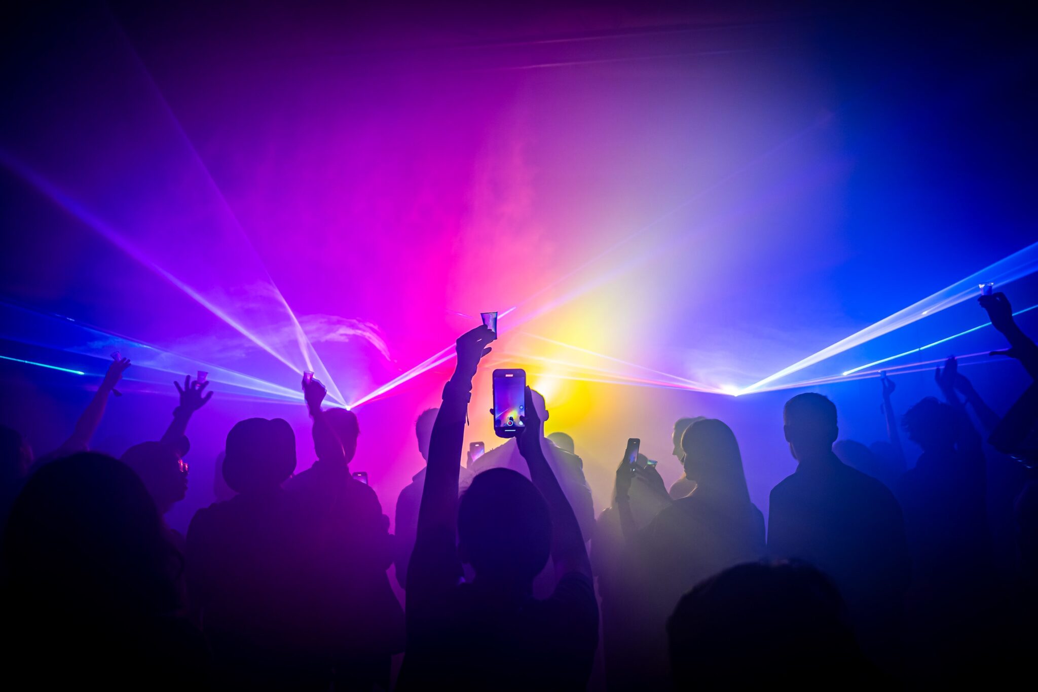 Silhouetted guests raising phones and hands in pink, violet and yellow haze.
