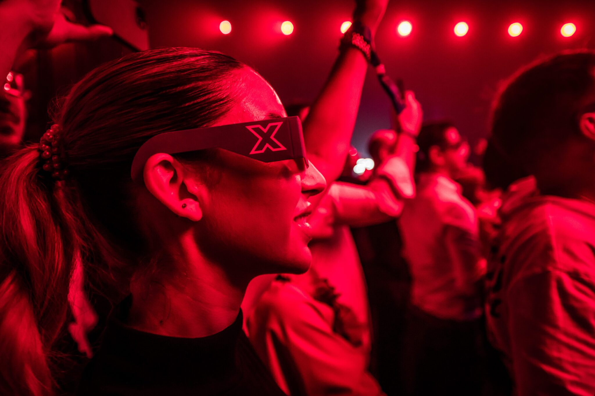Woman wearing futuristic glasses dancing in a crowd bathed in deep red light.