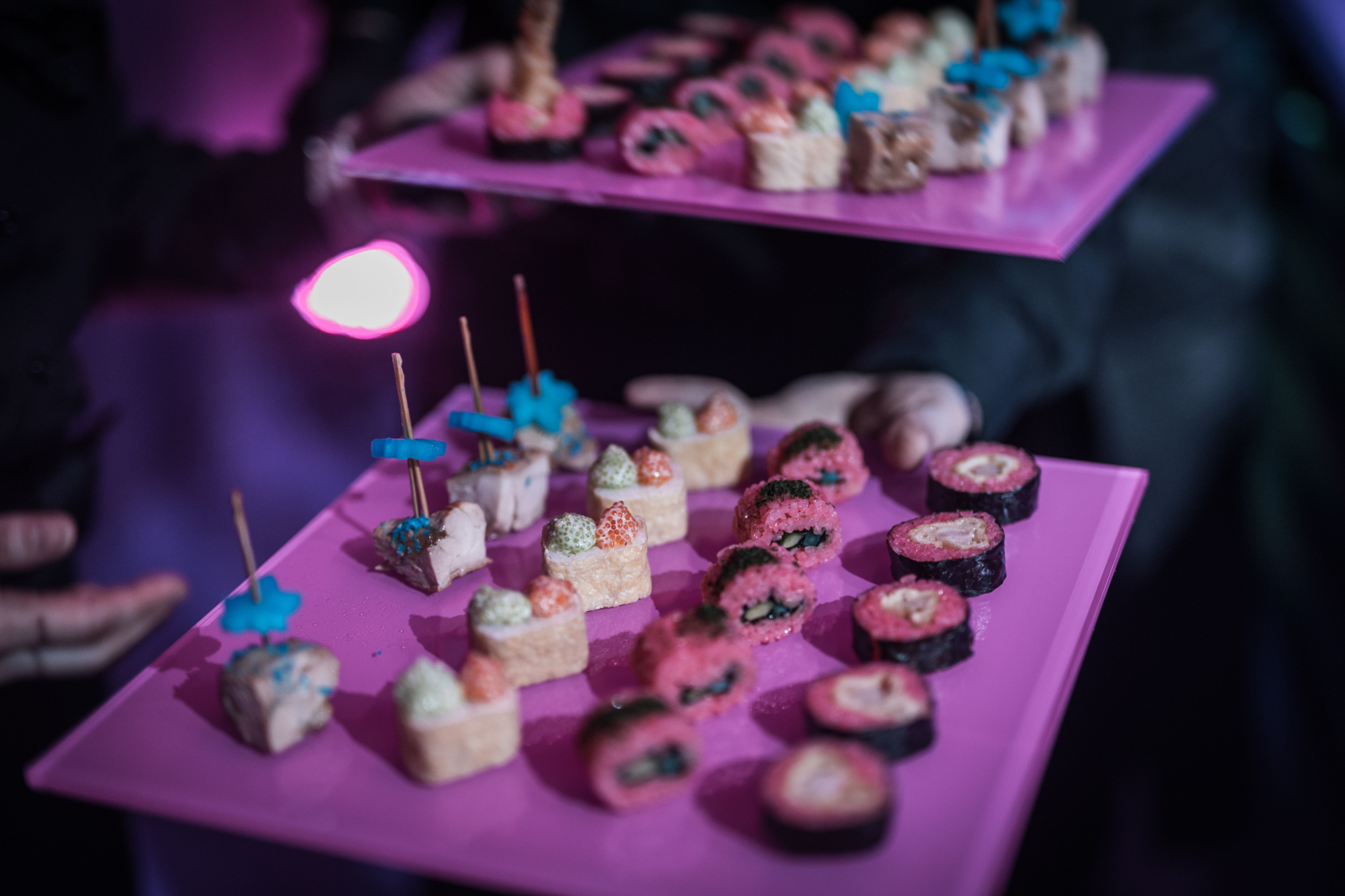 Pink trays of bite-sized sushi and canapés topped with colourful garnishes, under soft pink light.