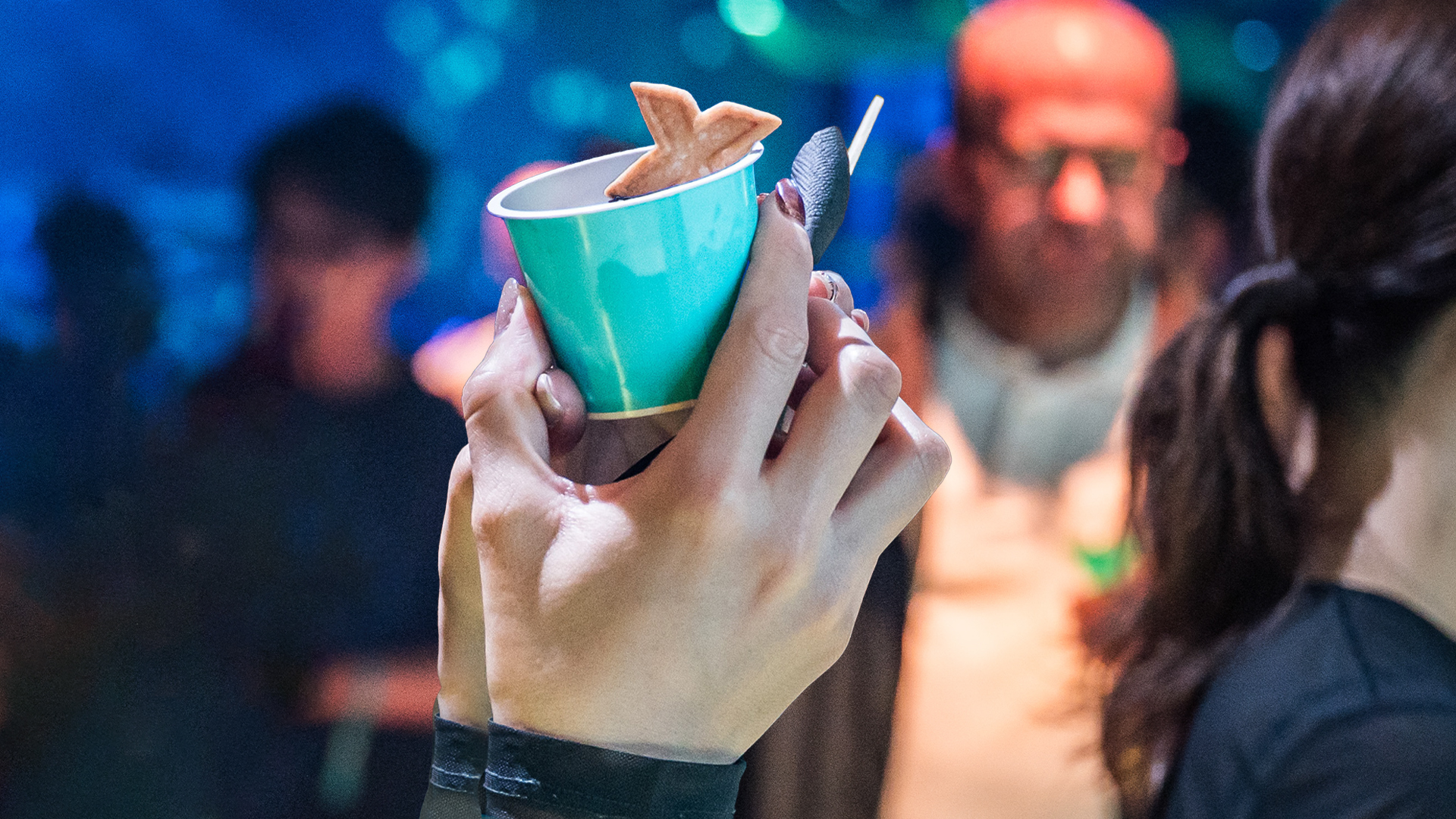 Close-up of a hand holding a small teal cup with a dessert, with blurred guests behind in blue light.