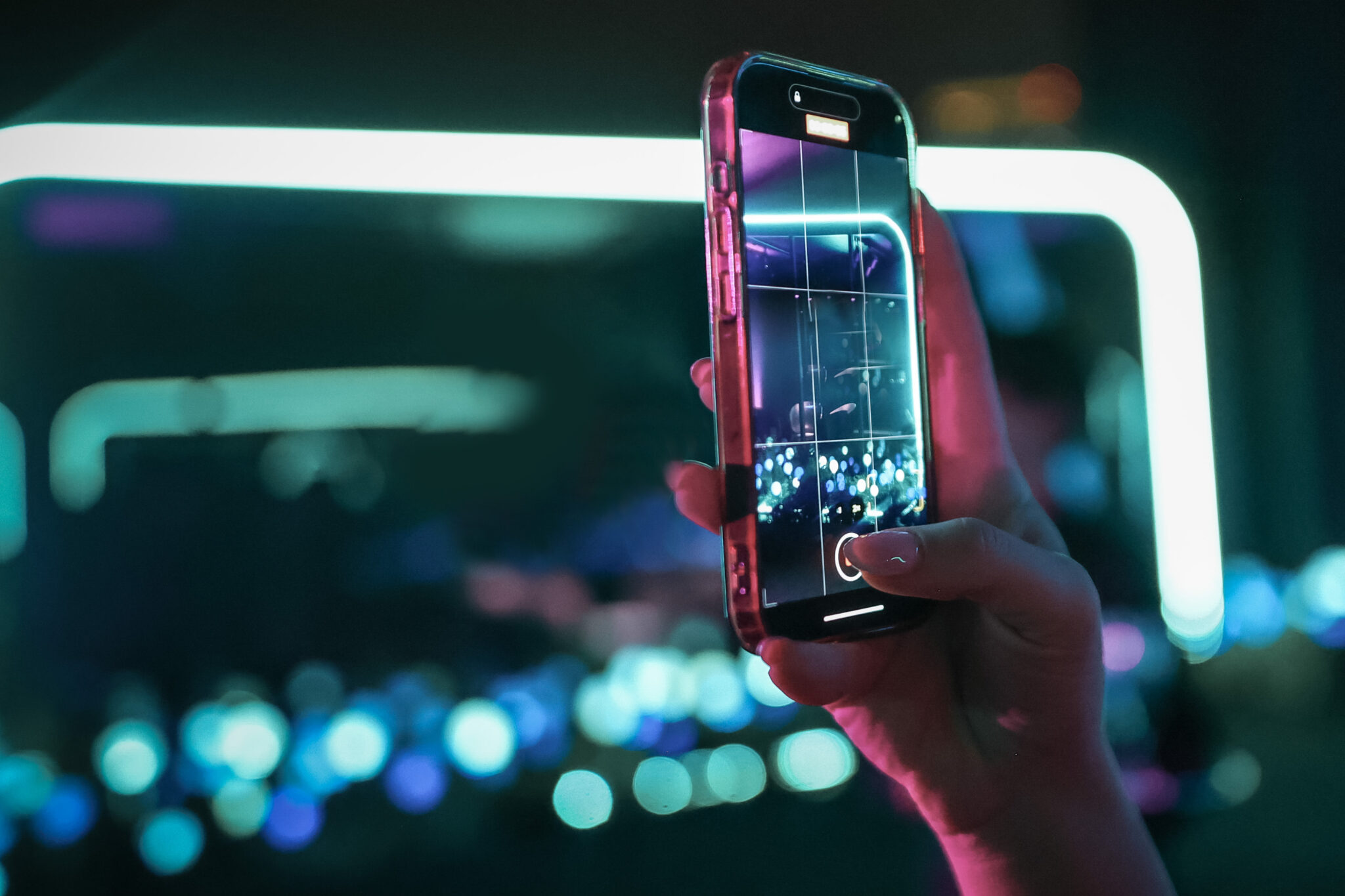 Hand holding a smartphone up to photograph a neon-lit outdoor garden with glowing orbs at night.