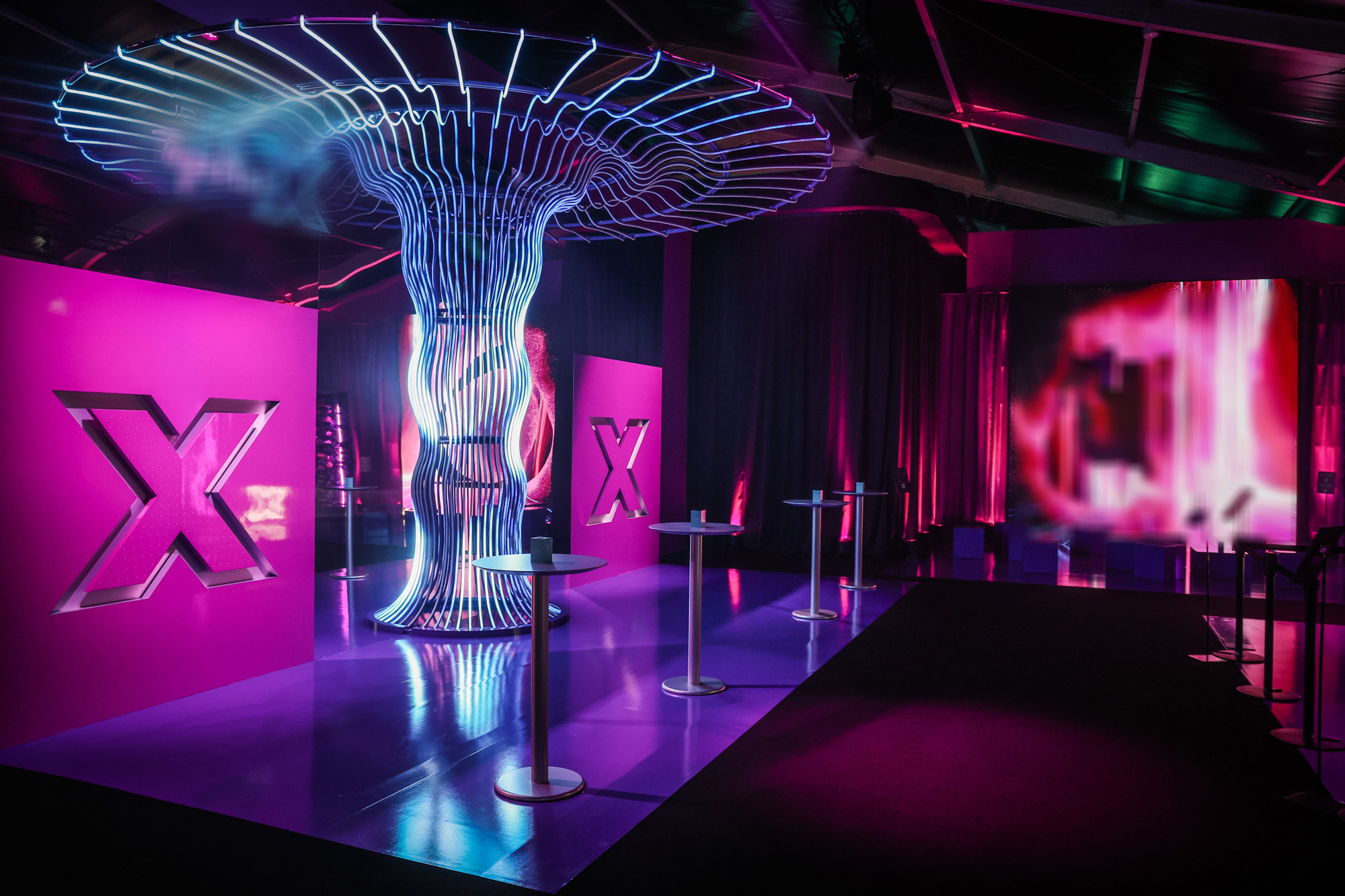 Empty zone with a tall wireframe tree-like light sculpture between pink illuminated panels and cocktail tables.