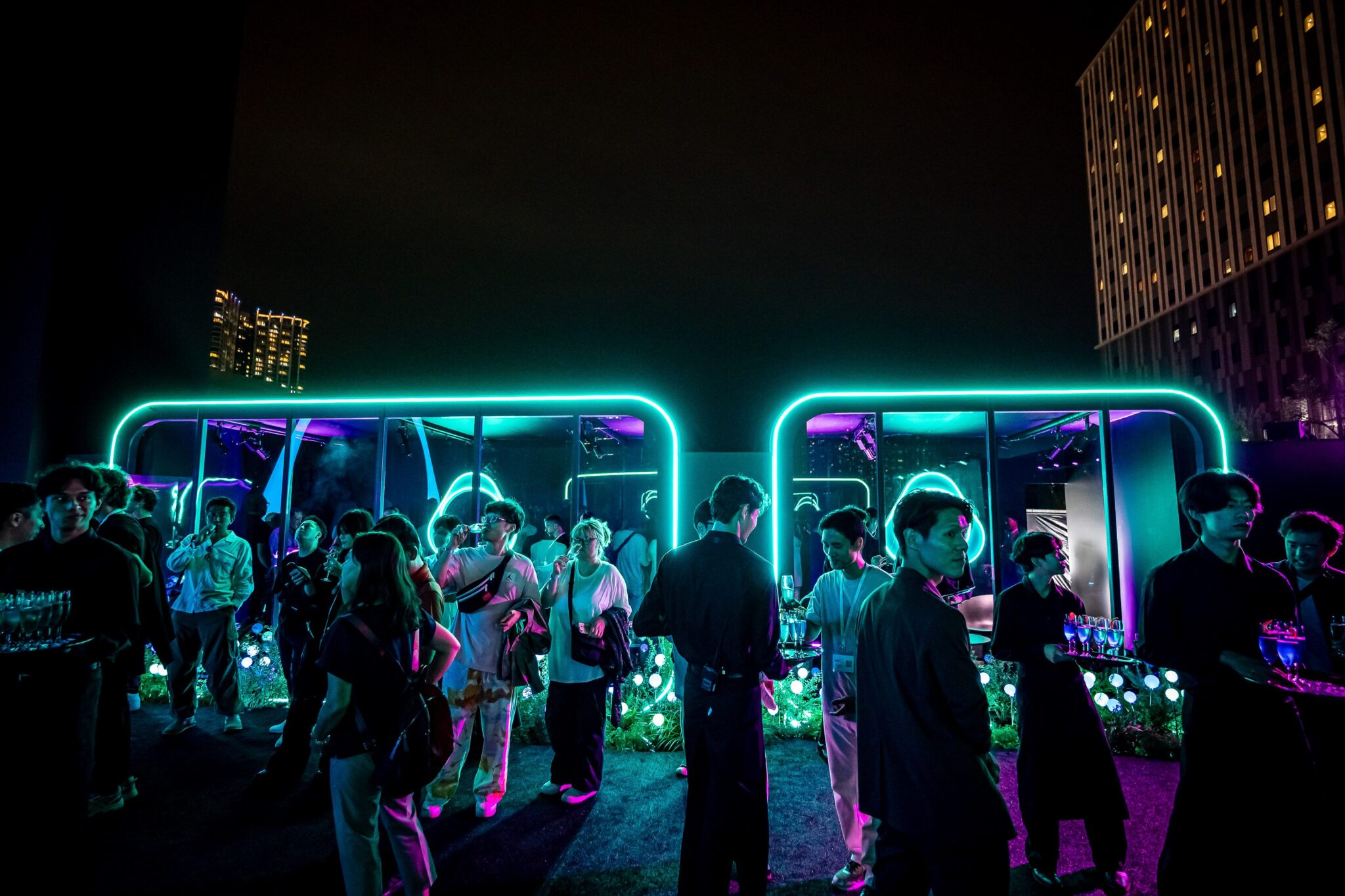 Guests walking past glowing neon-outlined pod structures in an outdoor garden at night, with a tall building behind.