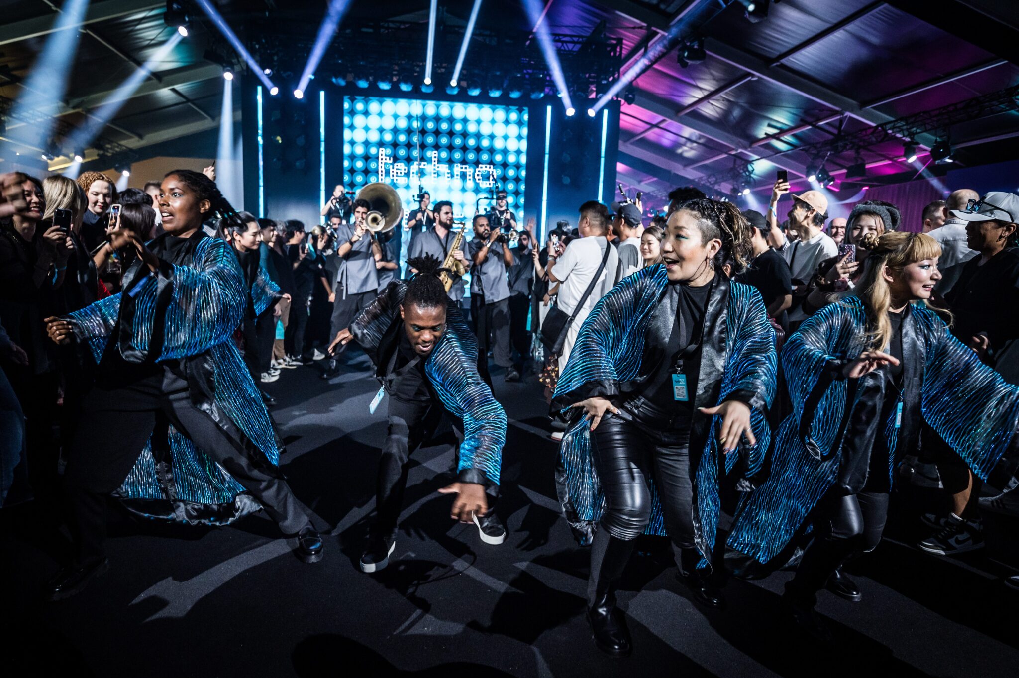 Dancers in dark-blue jackets performing energetically in front of a stage with a bright LED screen.