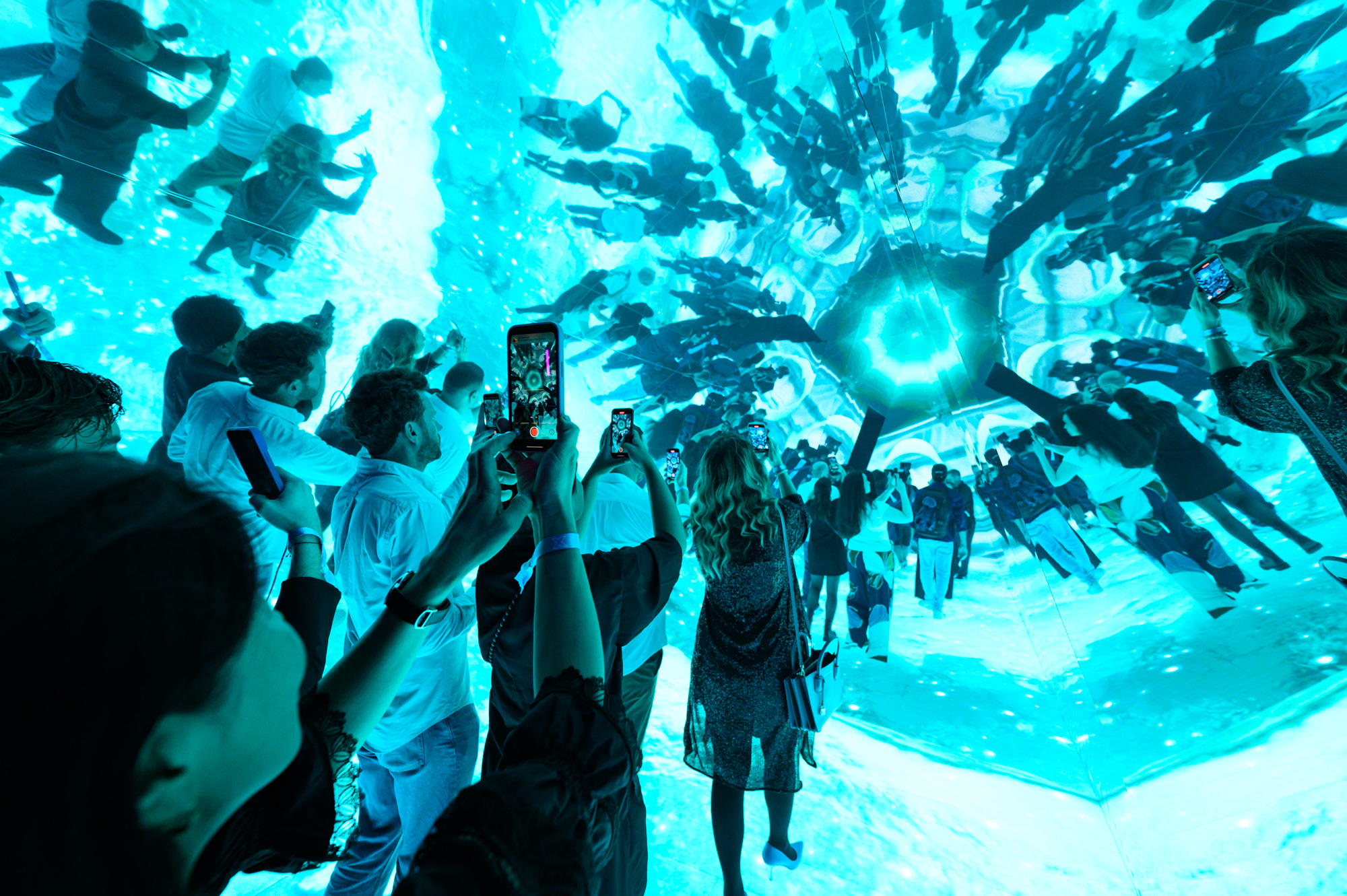 Guests filming a vivid turquoise immersive installation with branching light patterns overhead.