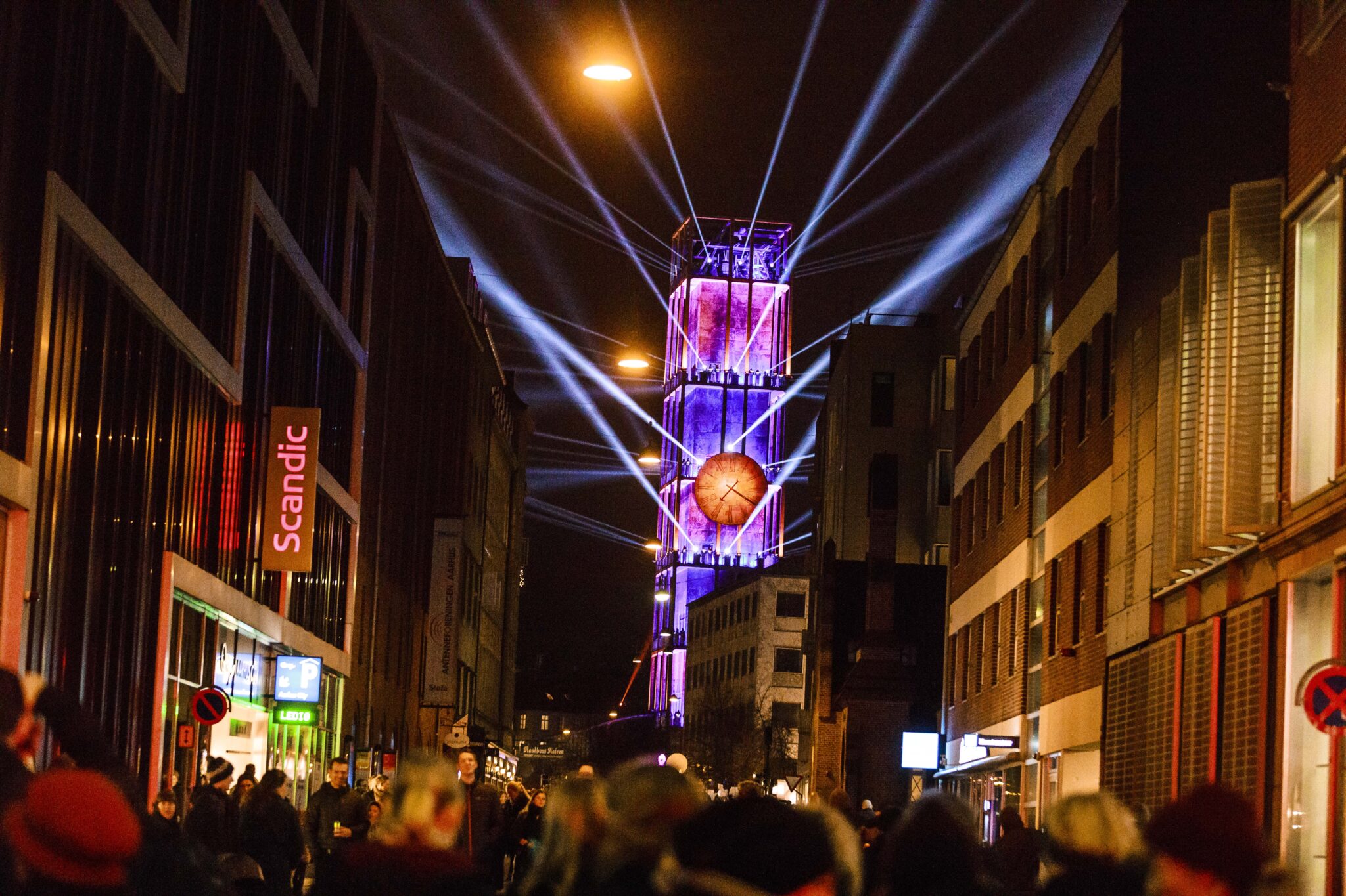 A crowd gathers in a city street at night as powerful beams of light radiate from a clock tower lit in purple and red hues. Battle Royal Studios