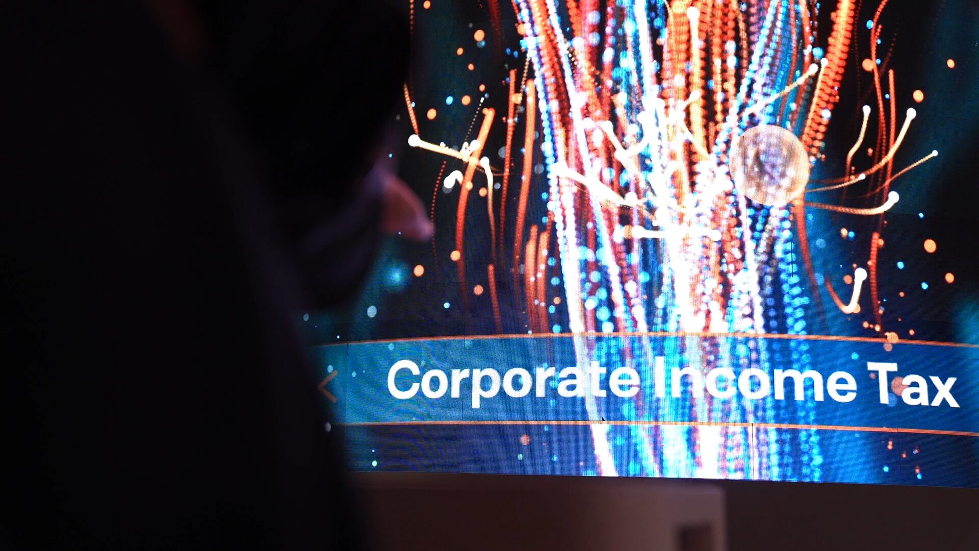 Close-up of an LED tower screen showing orange and blue streams of light with a banner reading Corporate Income Tax, with a visitor's silhouette in the foreground.