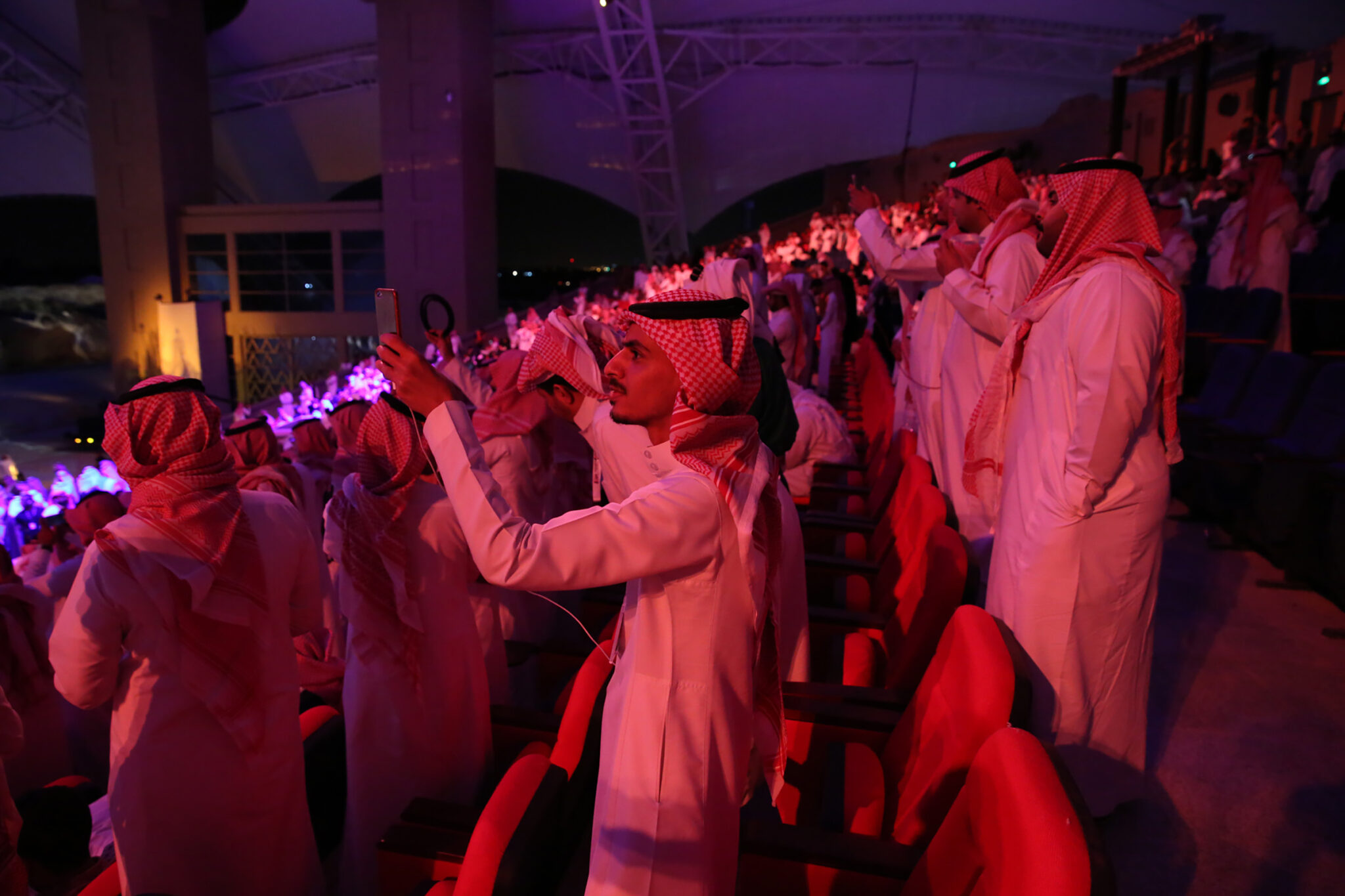 A crowd photographed from behind in deep red light, many holding up phones towards a stage out of frame.