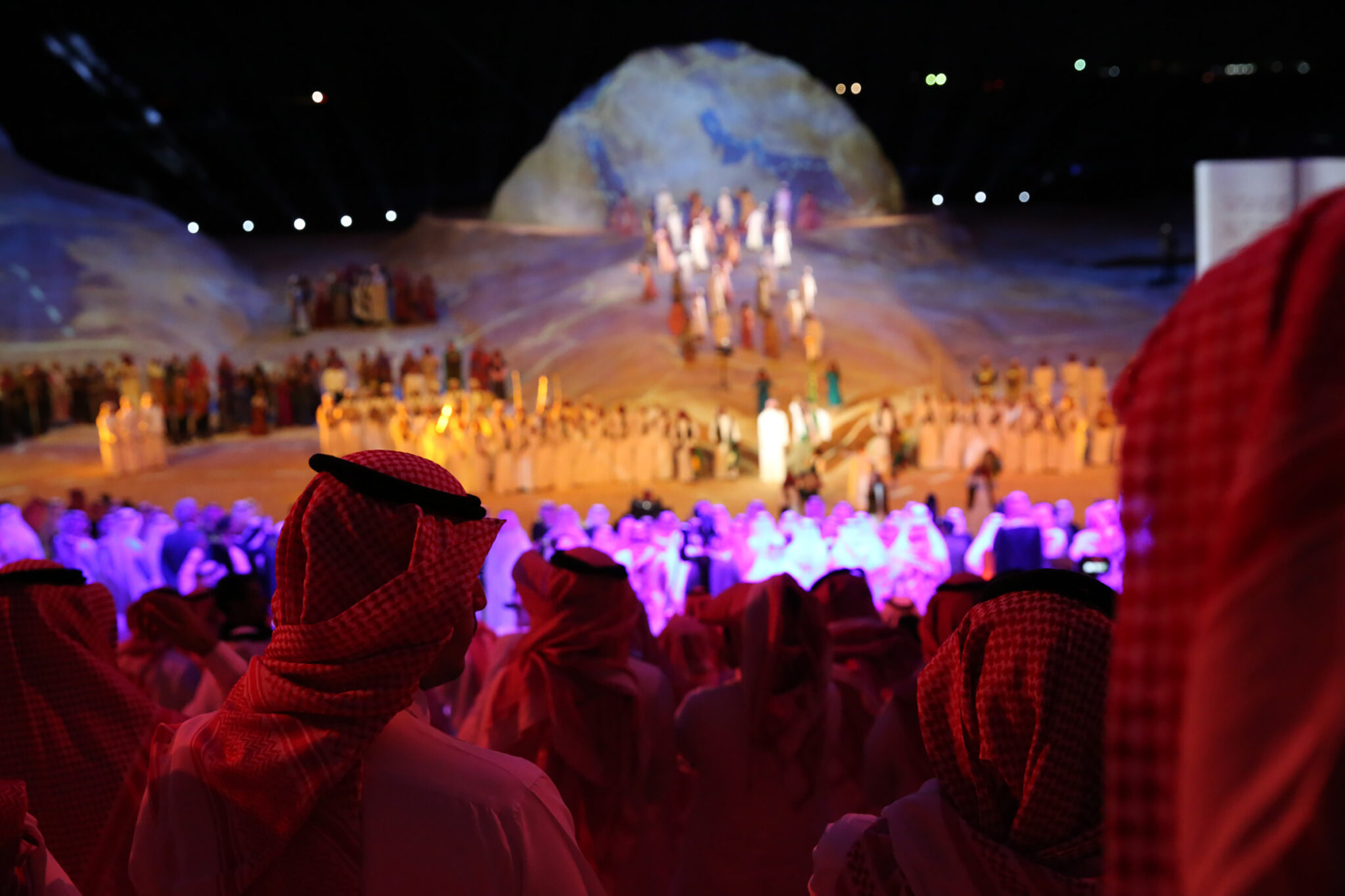 An audience in red-lit foreground silhouette watching a brightly lit stage with fireworks rising behind a rock-shaped set.