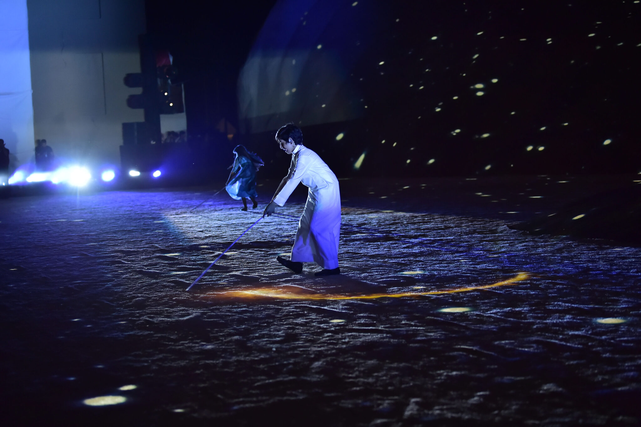 A lone performer crouched on a sand-covered stage in a single beam of light, surrounded by darkness.