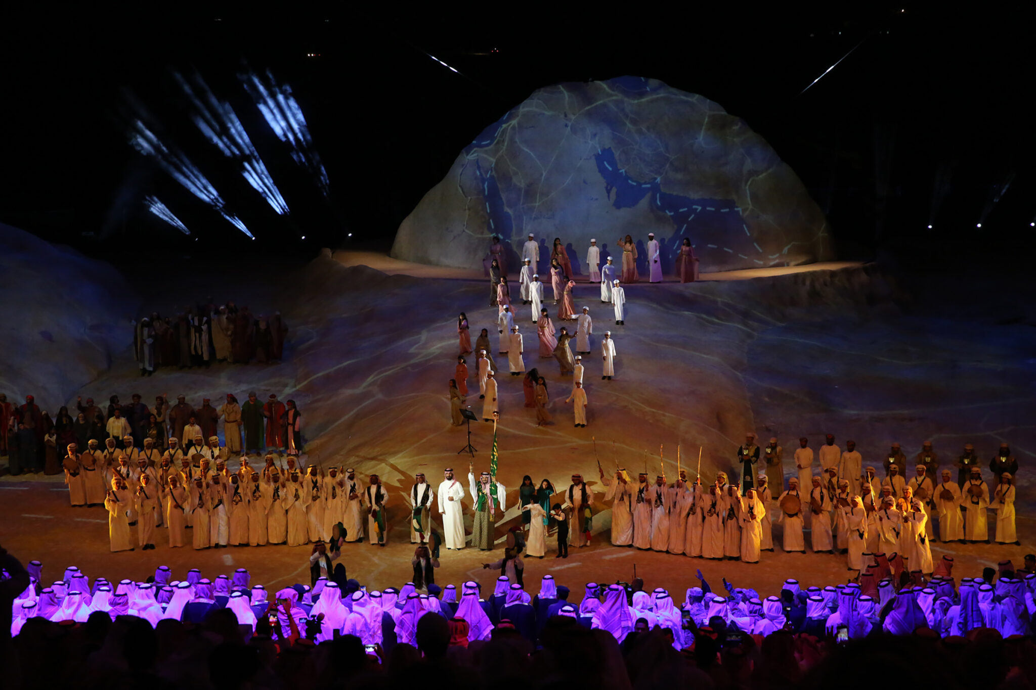 A large dome-shaped rock set carrying a projected map, rows of performers in traditional dress arranged across the stage below it, and an audience lit in purple in the foreground.