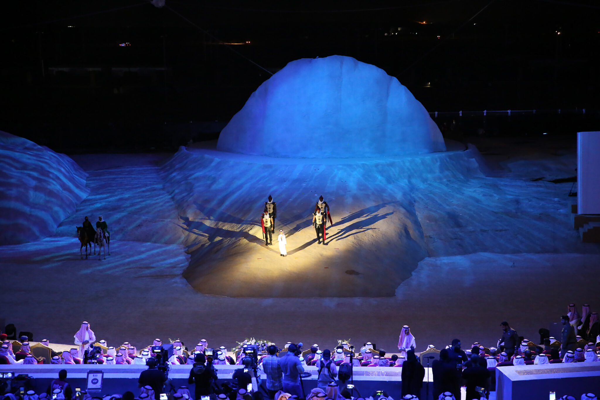 A wide stage built as a rocky outcrop, lit in deep blue, with a small boat and figures at its base and an orchestra in front.