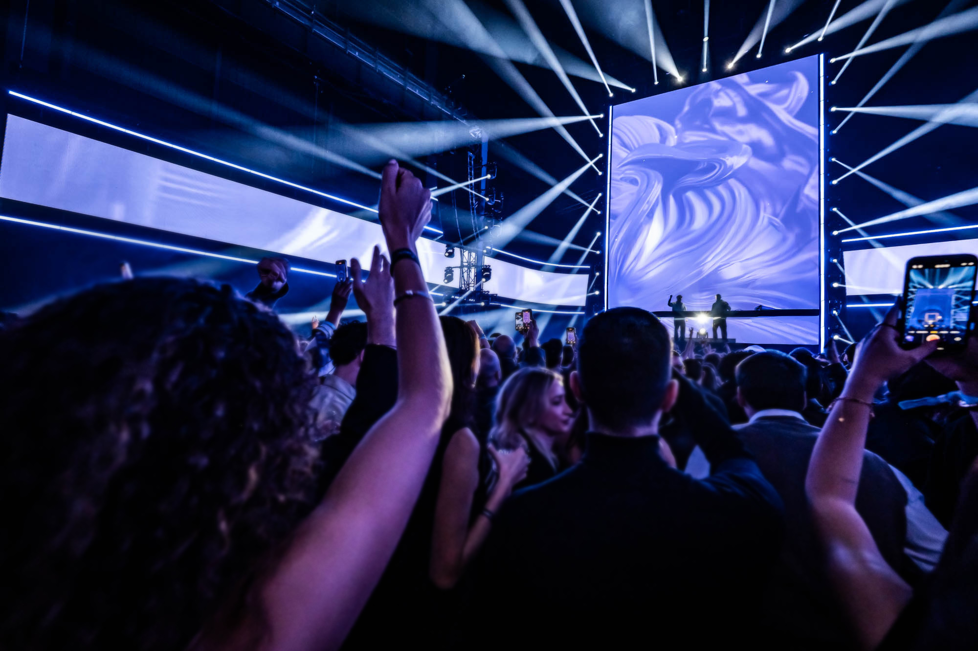 Fans raising their hands towards a stage screen showing blue swirling visuals.