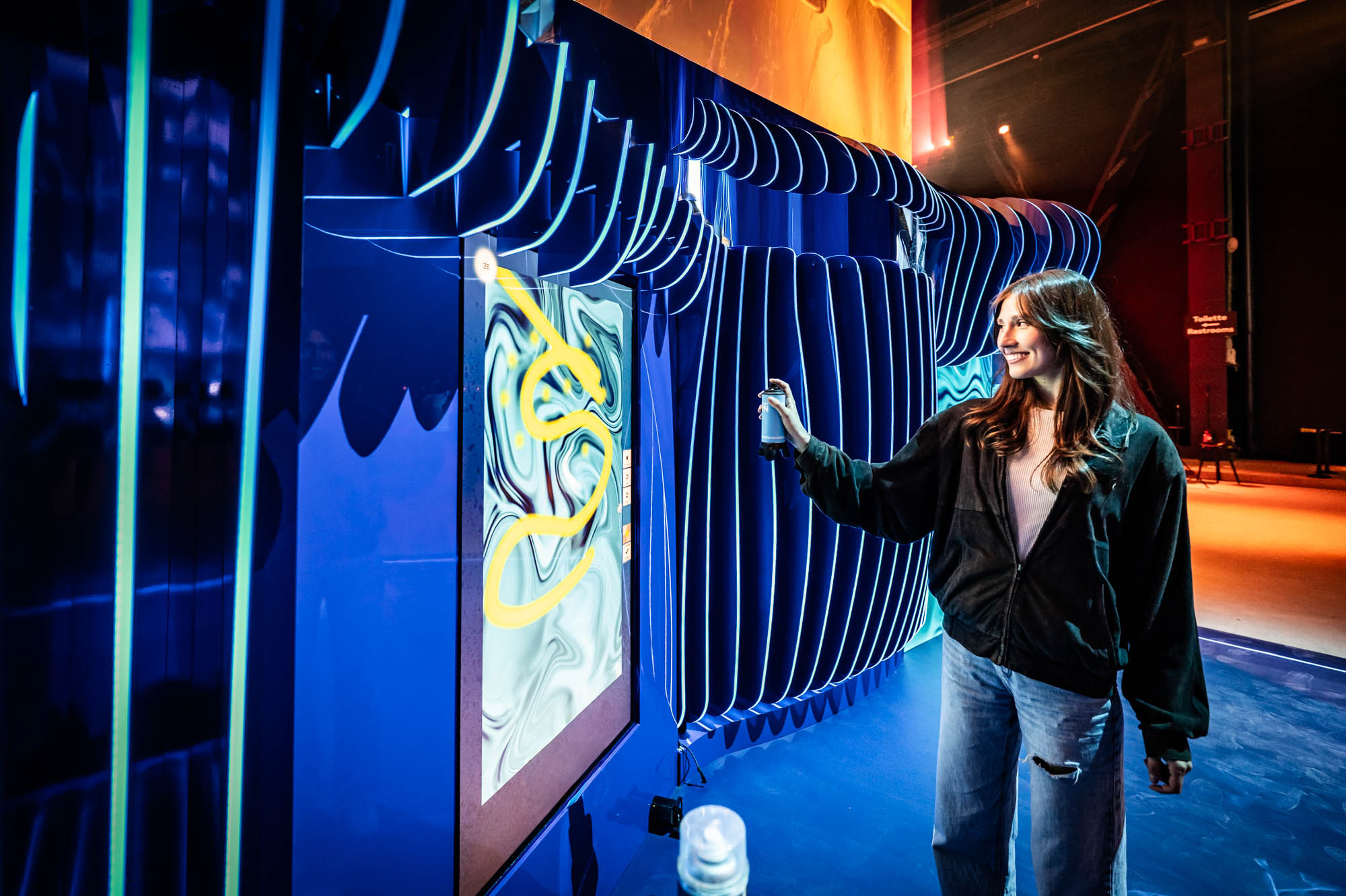 Woman in a dark jacket interacting with a screen showing a yellow drawing inside a blue ribbed installation.
