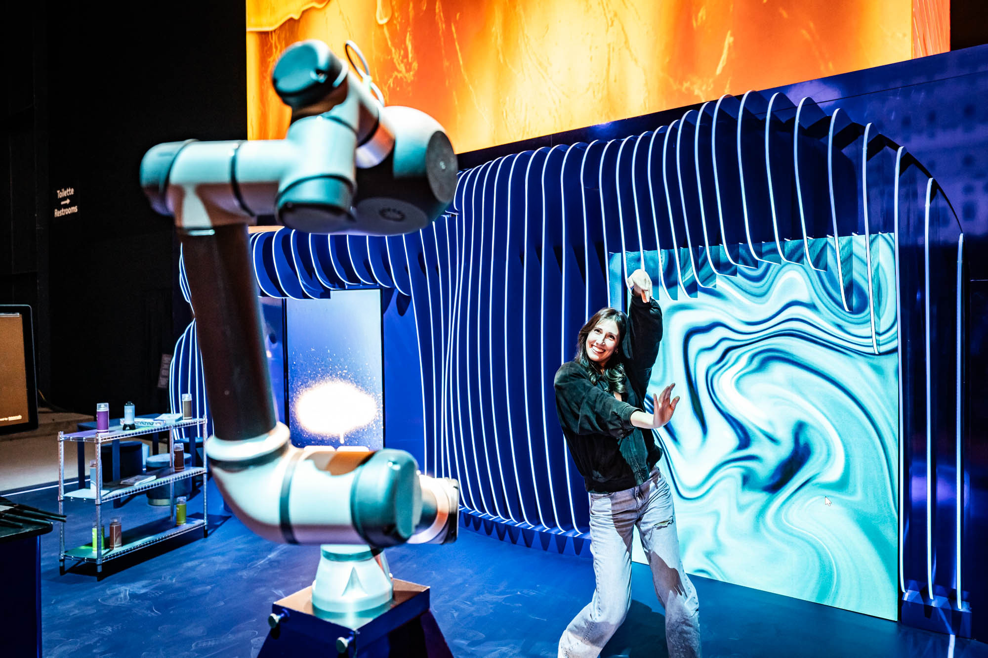 Woman touching a wall of swirling turquoise visuals next to a robotic arm on a blue stage.